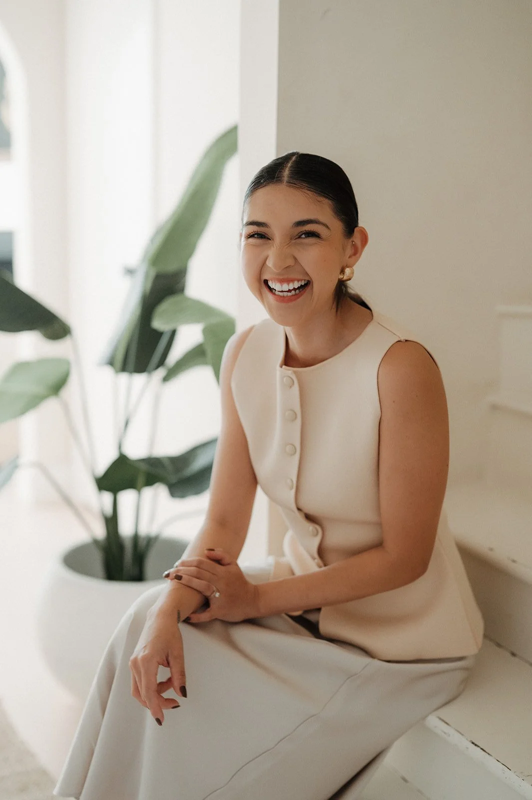 A woman in a cream-colored dress sitting on a white bench, smiling and laughing, with green houseplants in the background.