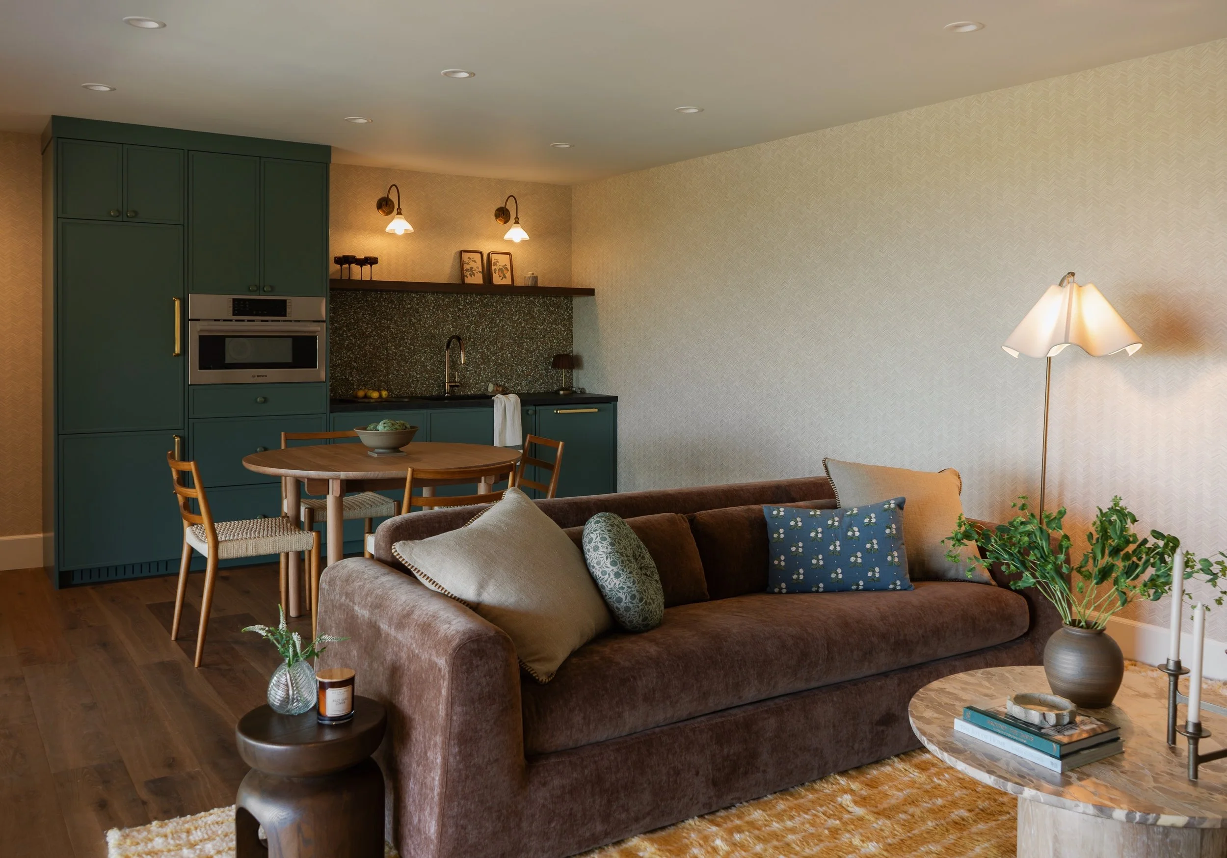 Living room with a brown sofa, decorative pillows, a marble side table with books and a plant, a round wooden coffee table with a candle and plant, a tan rug, green kitchen cabinets, a circular dining table with chairs, a kitchen sink, and wall-mounted lights.