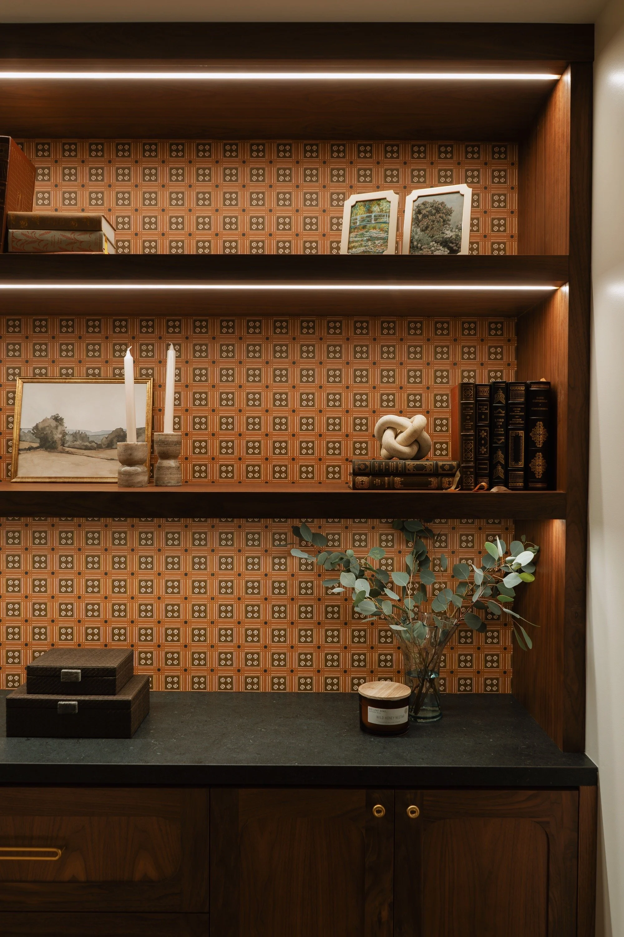 A wooden display shelf with two levels, decorated with framed artwork, candles, books, a knot sculpture, and a vase with eucalyptus branches against a patterned tile background.