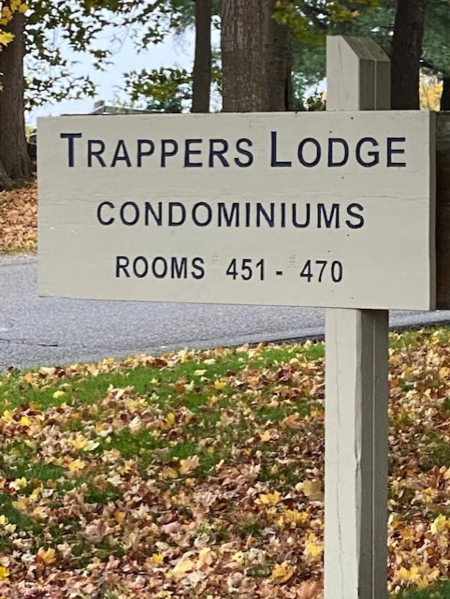 Sign for Trappers Lodge Condominiums, indicating rooms 451 through 470, outdoors amidst fallen autumn leaves and trees.
