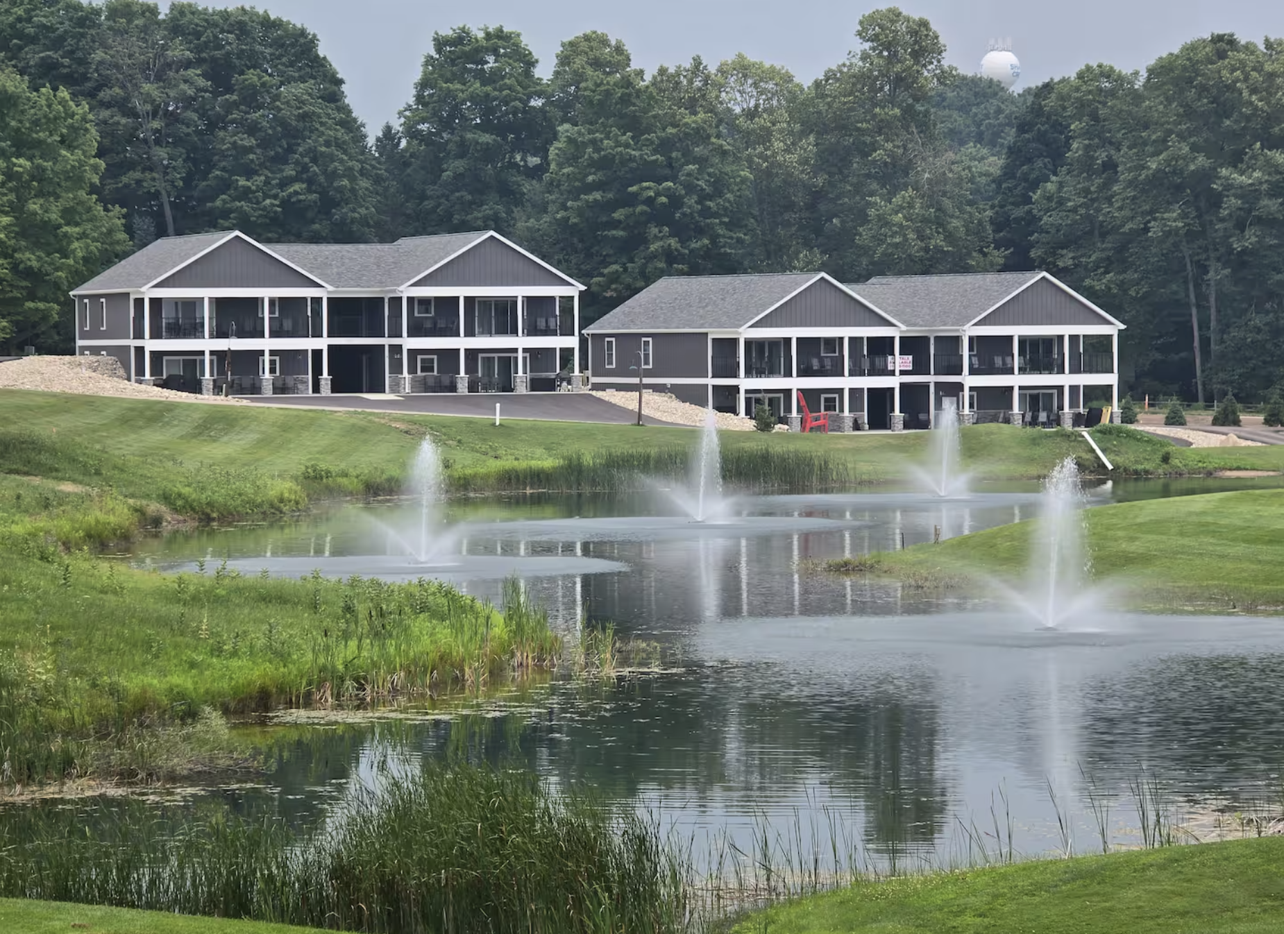 A scenic landscape featuring a calm lake with trees along the shoreline, dark silhouettes of trees and mountains in the background, and a colorful sunset sky with shades of yellow, orange, and blue.