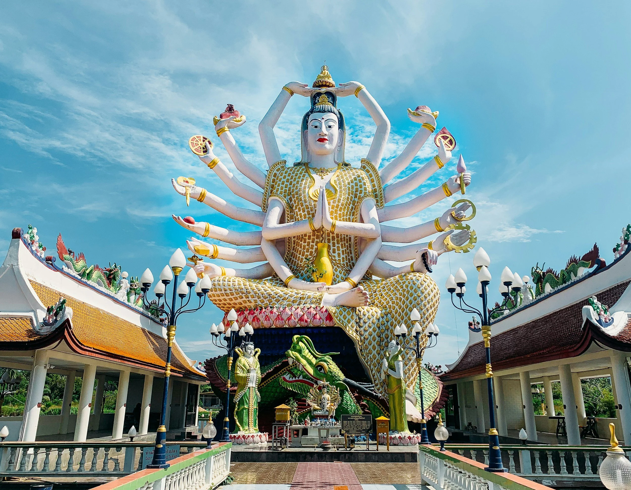 Large multi-armed statue of a deity sitting cross-legged outdoors, with a dragon dipped in green at the base and a blue sky background.