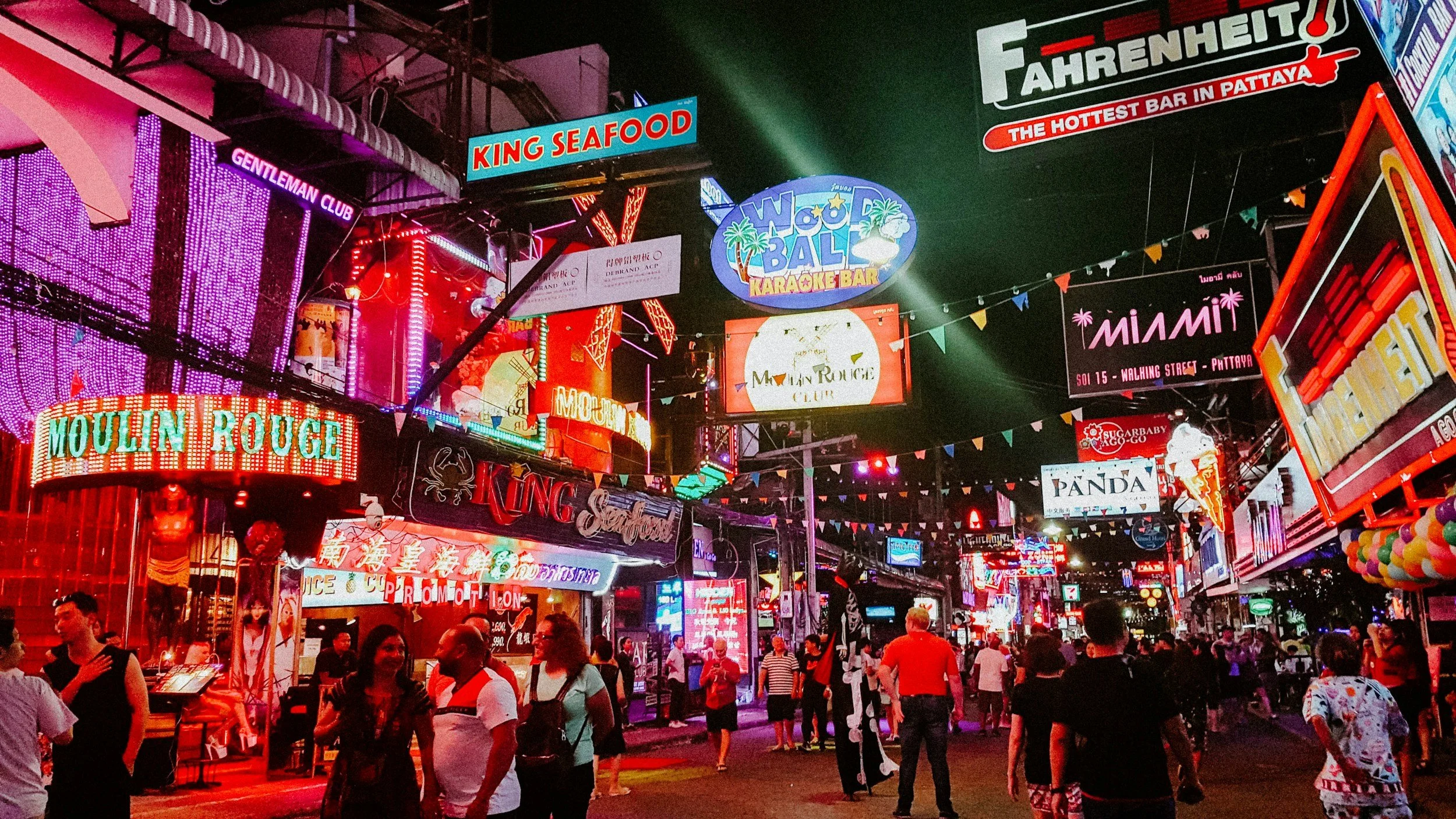 Night scene of a busy street filled with illuminated neon signs advertising bars, clubs, and eateries, with people walking and socializing.