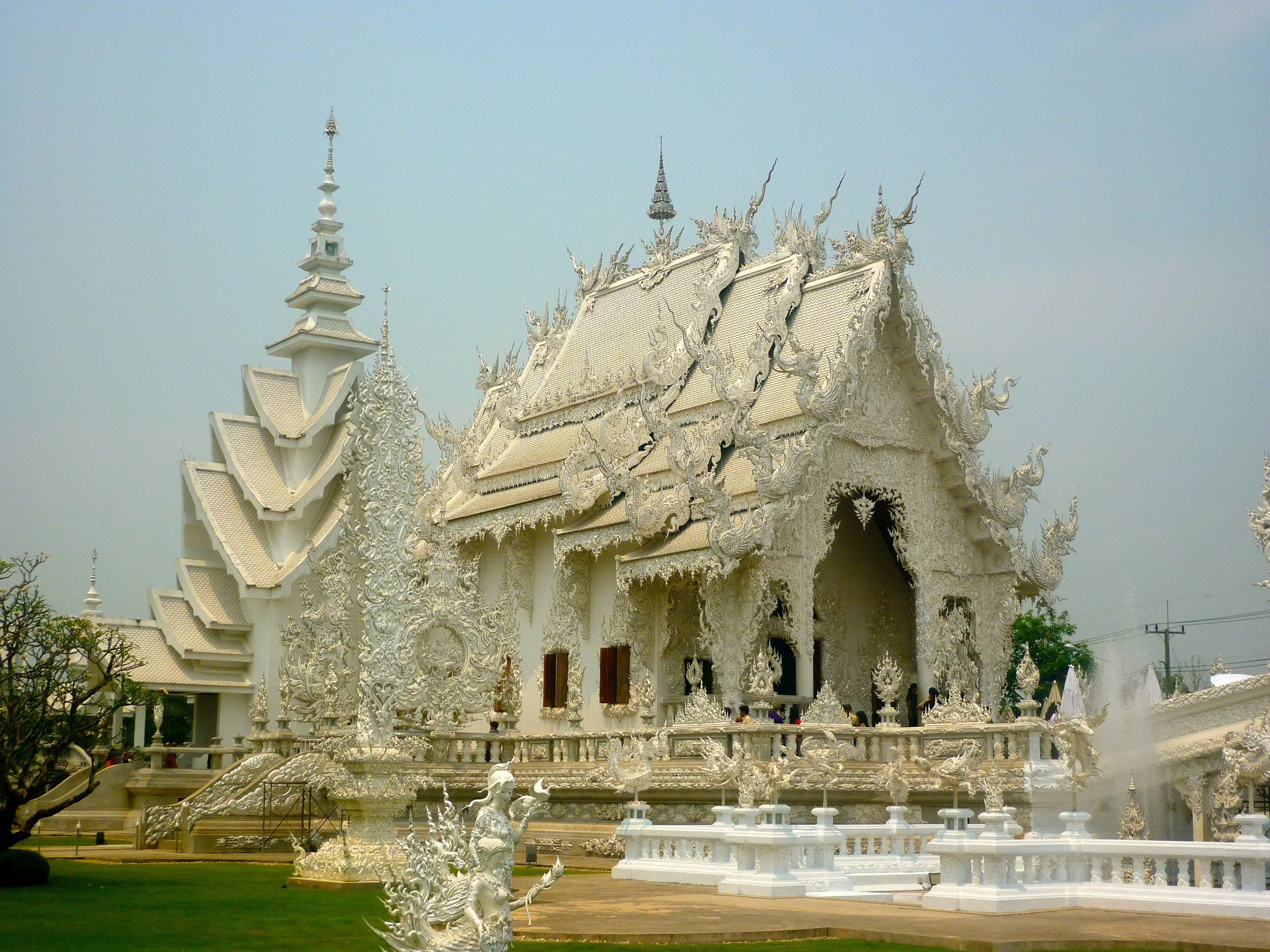White ornate temple with detailed carvings and multiple layered roof tiles, set against a cloudy sky, with some visitors visible near the entrance.