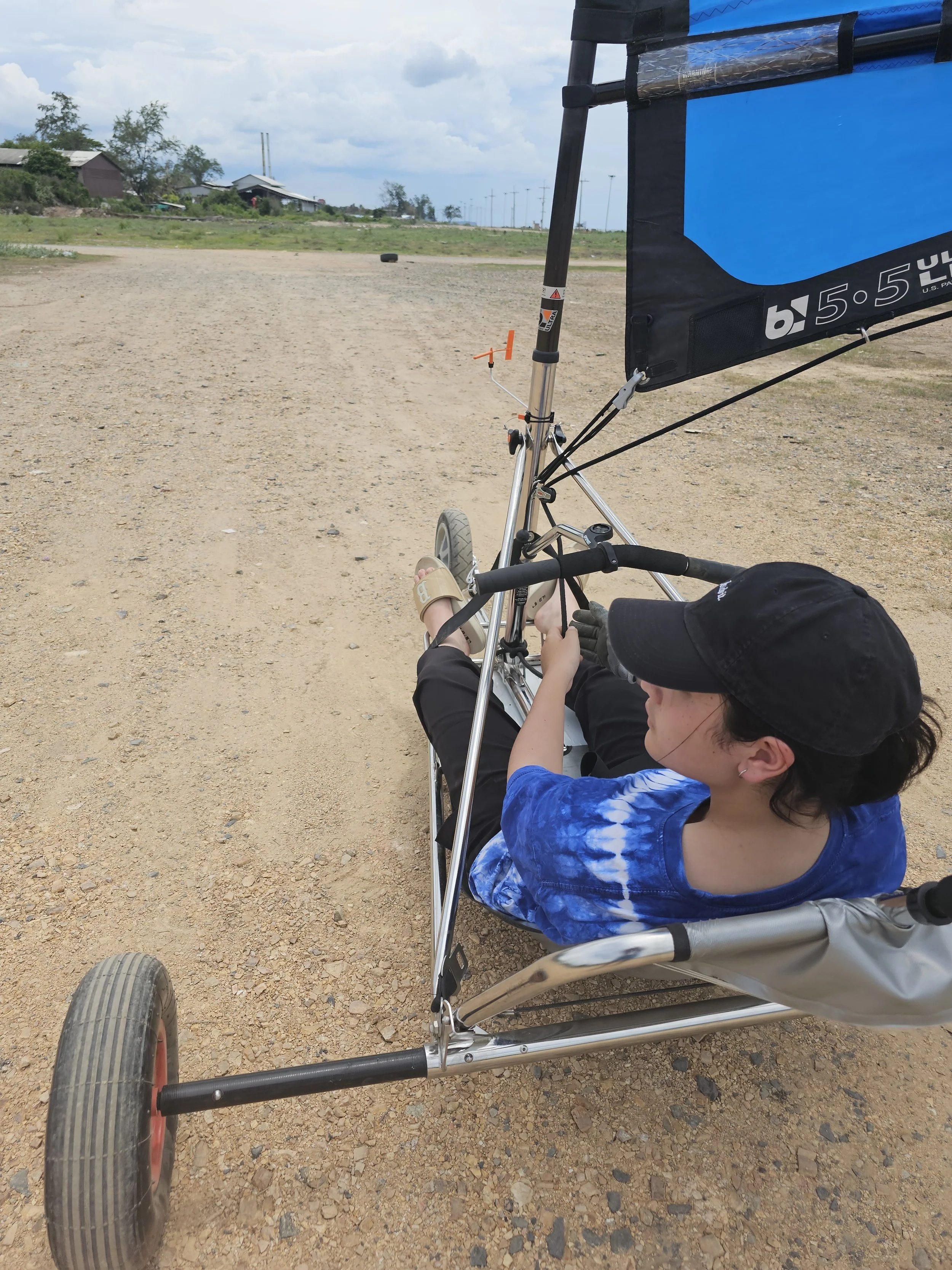A person sitting in a blue and black blokart on a dirt track, wearing a black cap, black shorts, a blue tie-dye shirt, and sandals, with a few houses and trees in the background under a partly cloudy sky.