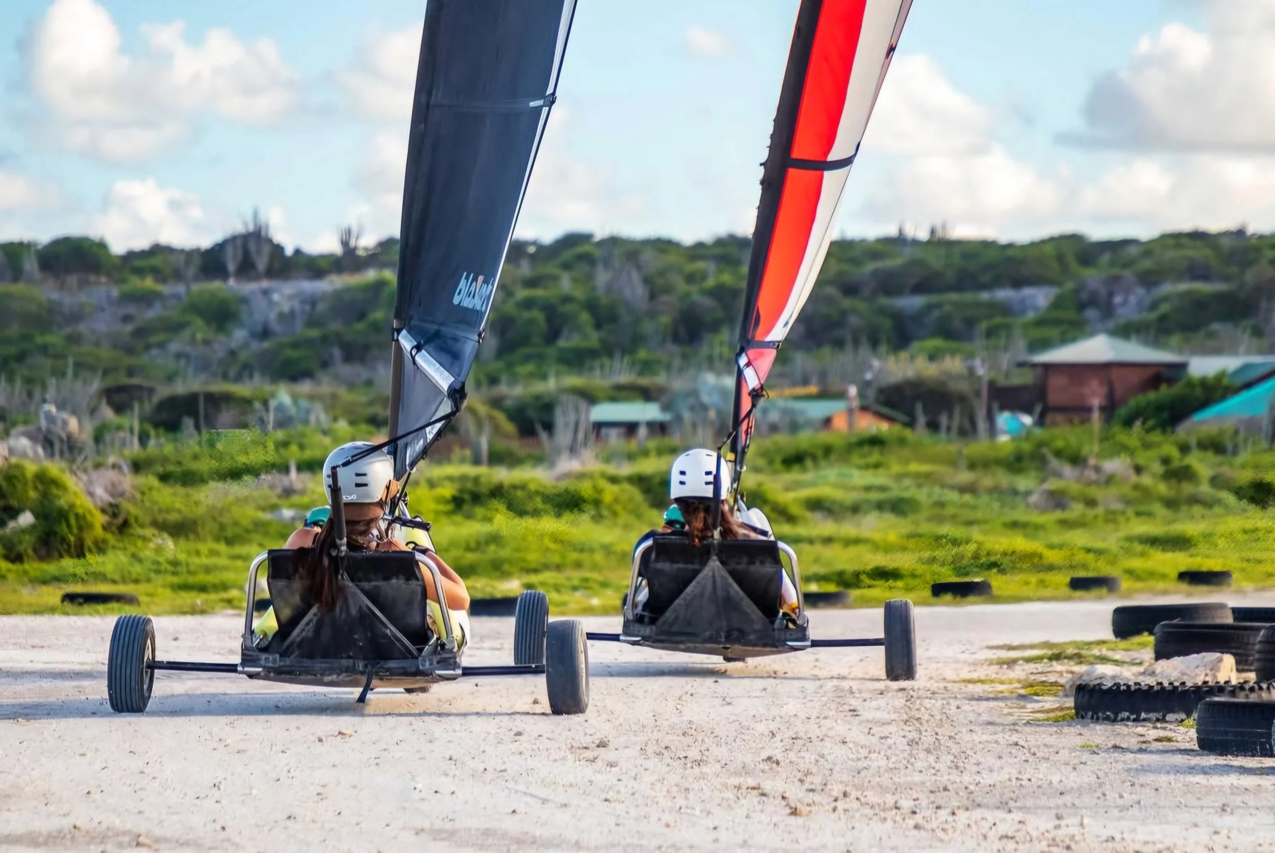 Two people riding on land sailboats with large sails on a sandy track surrounded by green landscape and tires used as obstacles.