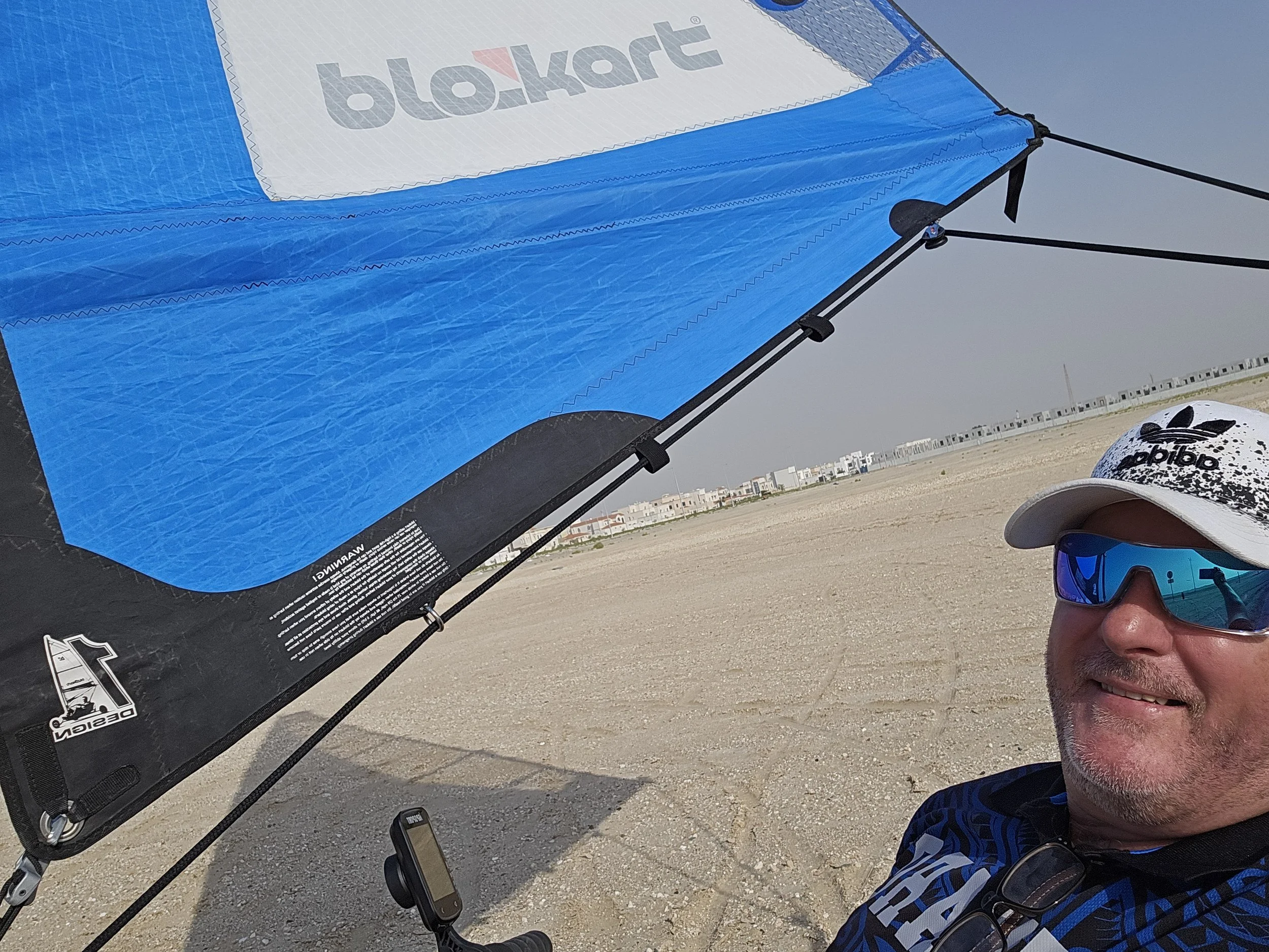A man wearing sunglasses and a white Adidas cap takes a selfie under a large  blokart  with the Blokart logo, on a sandy beach with a distant row of white buildings.