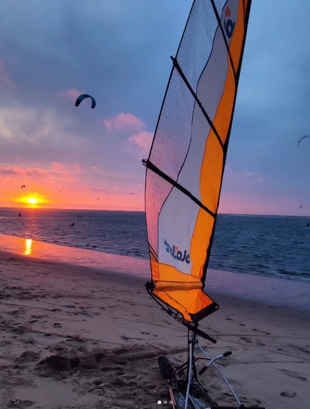 A beach at sunset with a sail, possibly part of a land yacht, and kite surfers in the distance over the water.