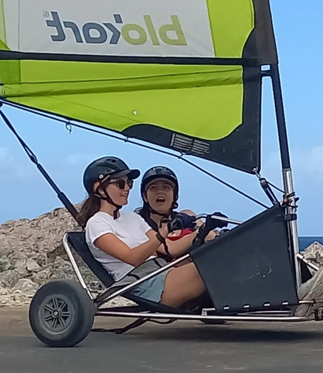 Two women wearing helmets and sunglasses riding in a two-person  blokart  with a sail attached, on a rocky shoreline with the ocean in the background.