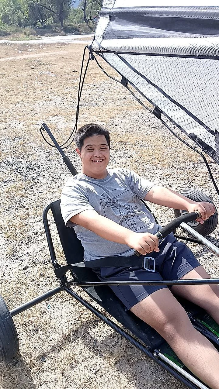 Boy smiling and sitting in a land sailing vehicle outdoors on a dirt surface.
