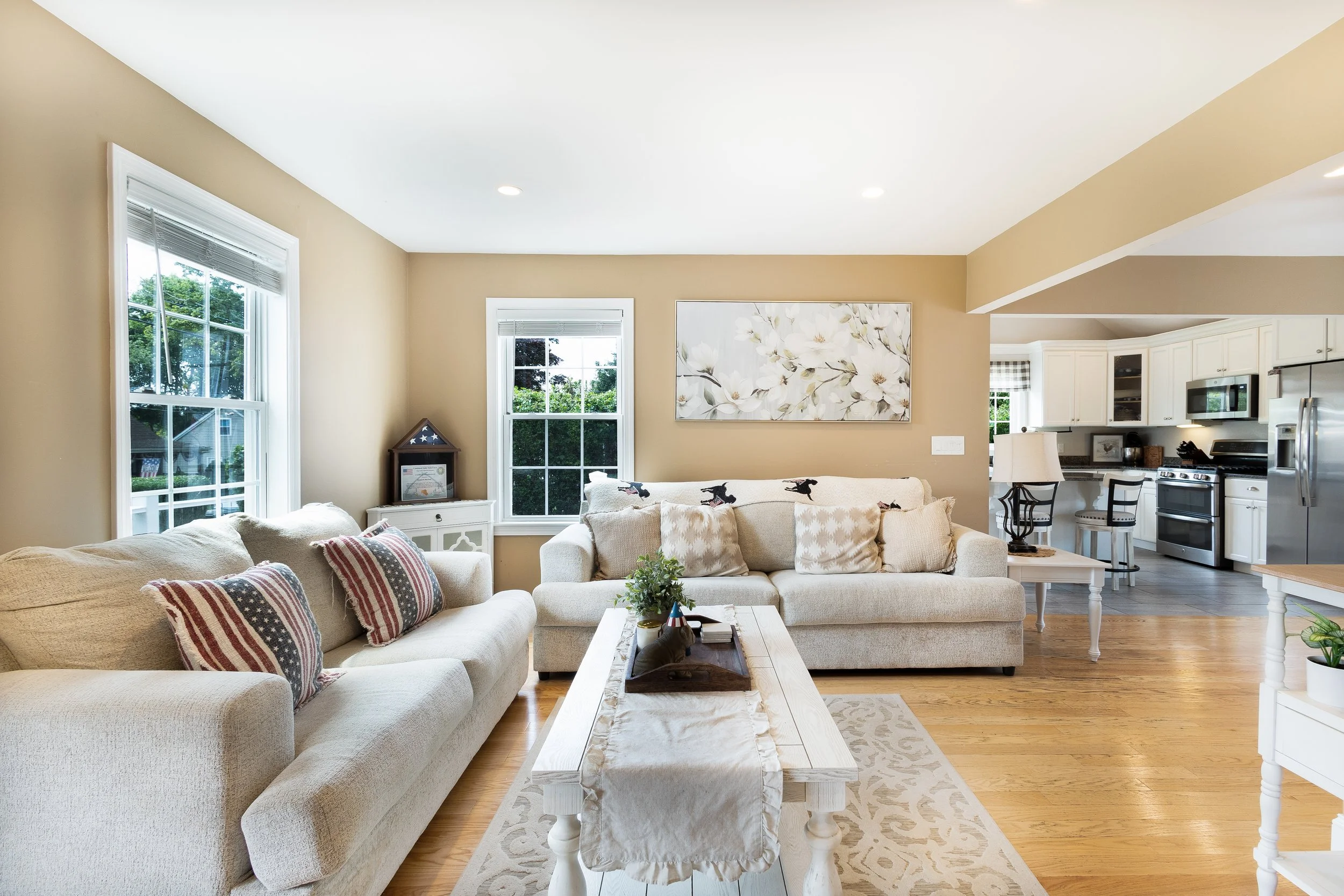 Bright living room with beige walls, white sofas, and hardwood floors, adjacent to a kitchen with white cabinets and stainless steel appliances.