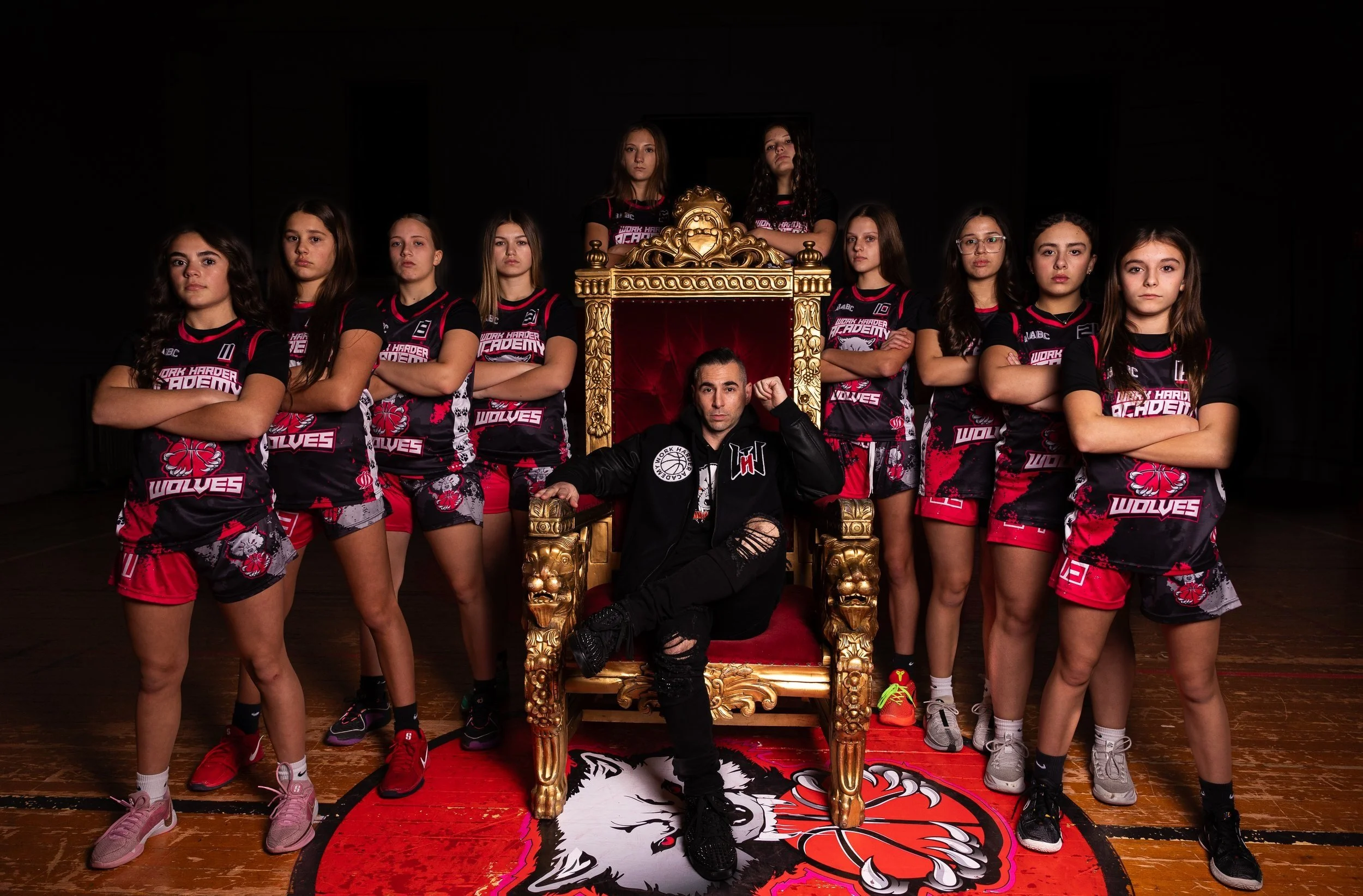 A men's basketball coach sitting on a throne-like chair surrounded by nine female basketball players on a court, with a team logo on the floor.