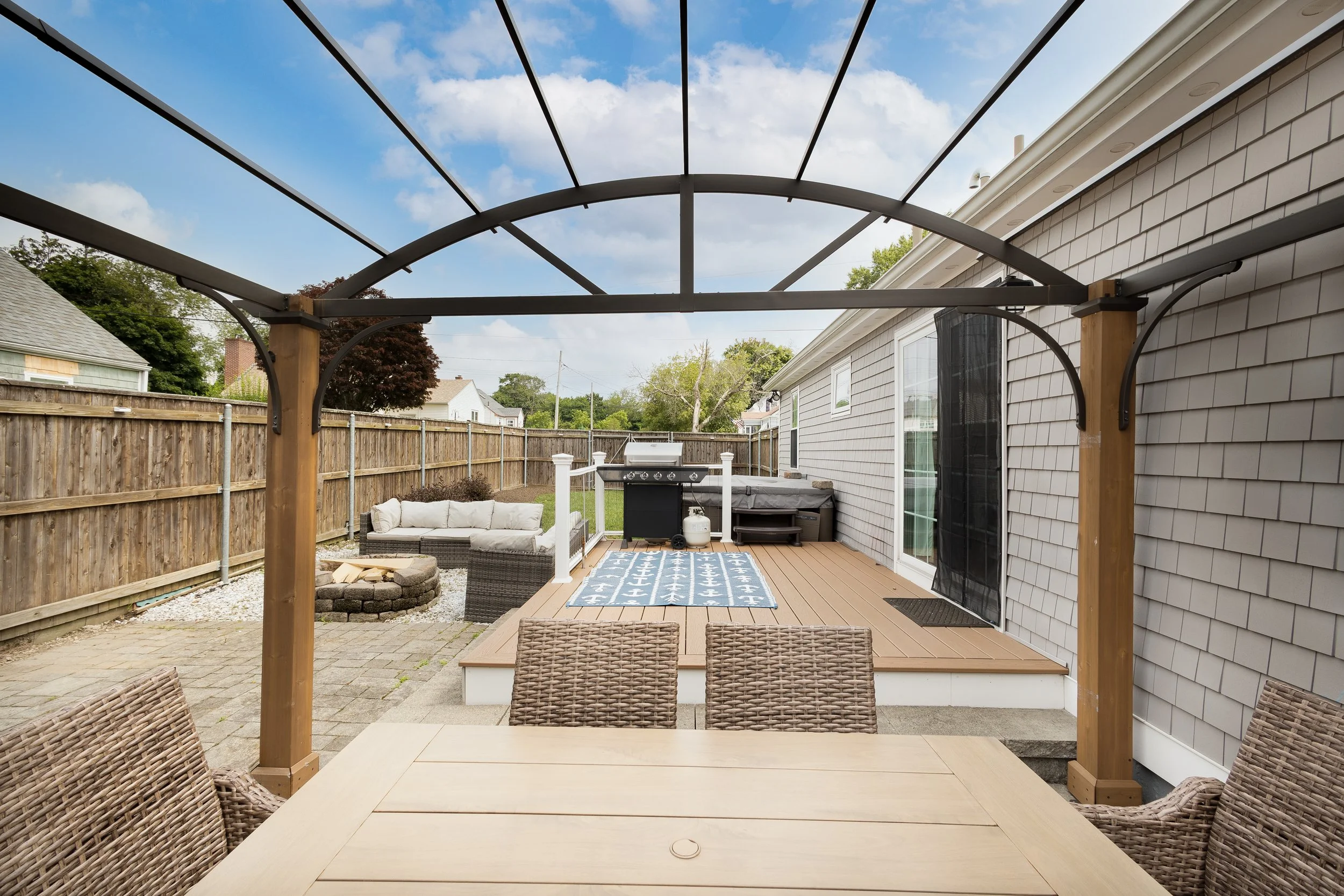 A backyard patio area with a dining table and chairs in the foreground, a wooden deck with a rug, a grill, hot tub, and outdoor seating, and a fire pit area with a garden couch in the background, surrounded by a wooden fence.