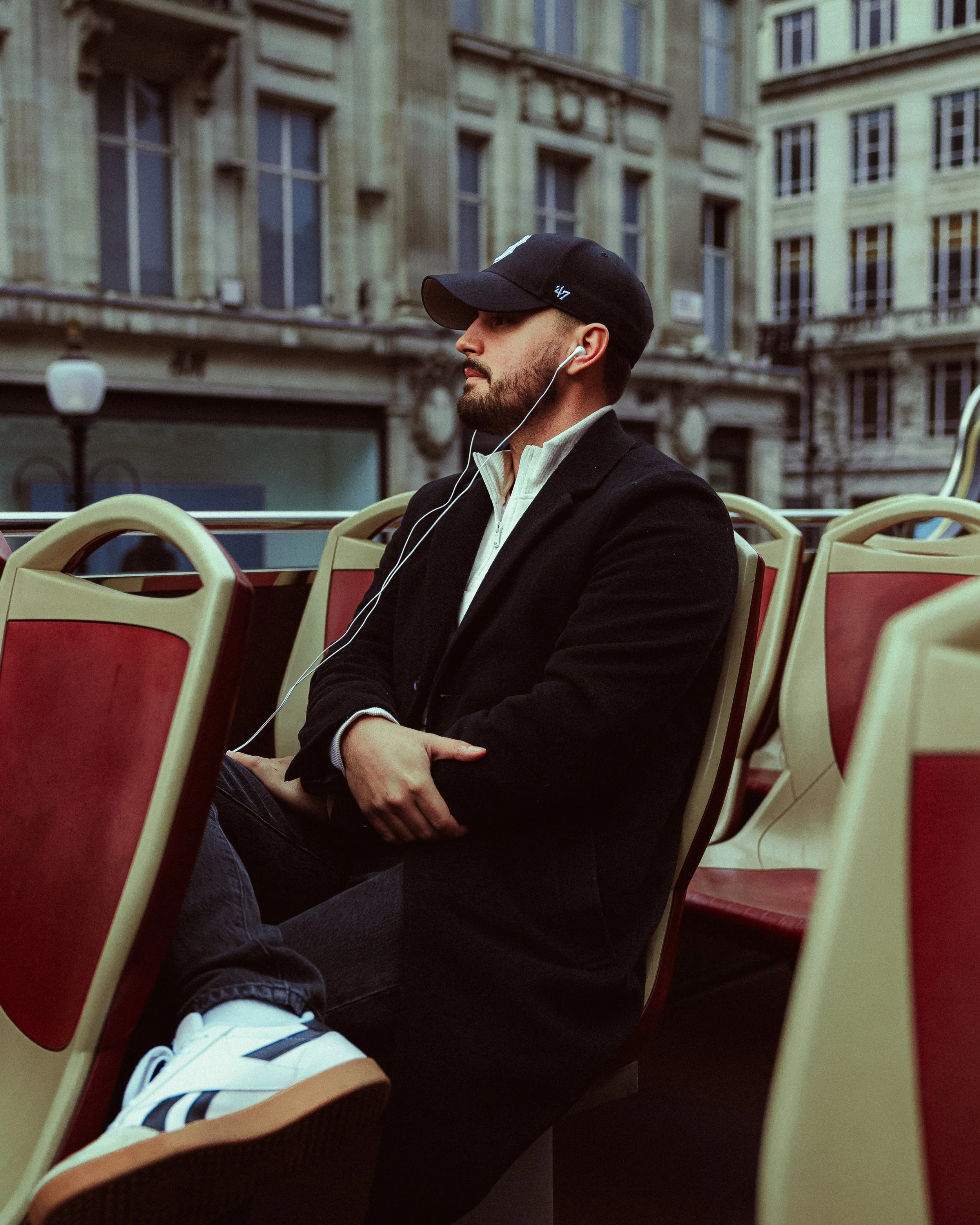 A man sitting on a red and beige bus seat wearing headphones, a black cap, a black jacket, and sneakers, with urban buildings in the background.