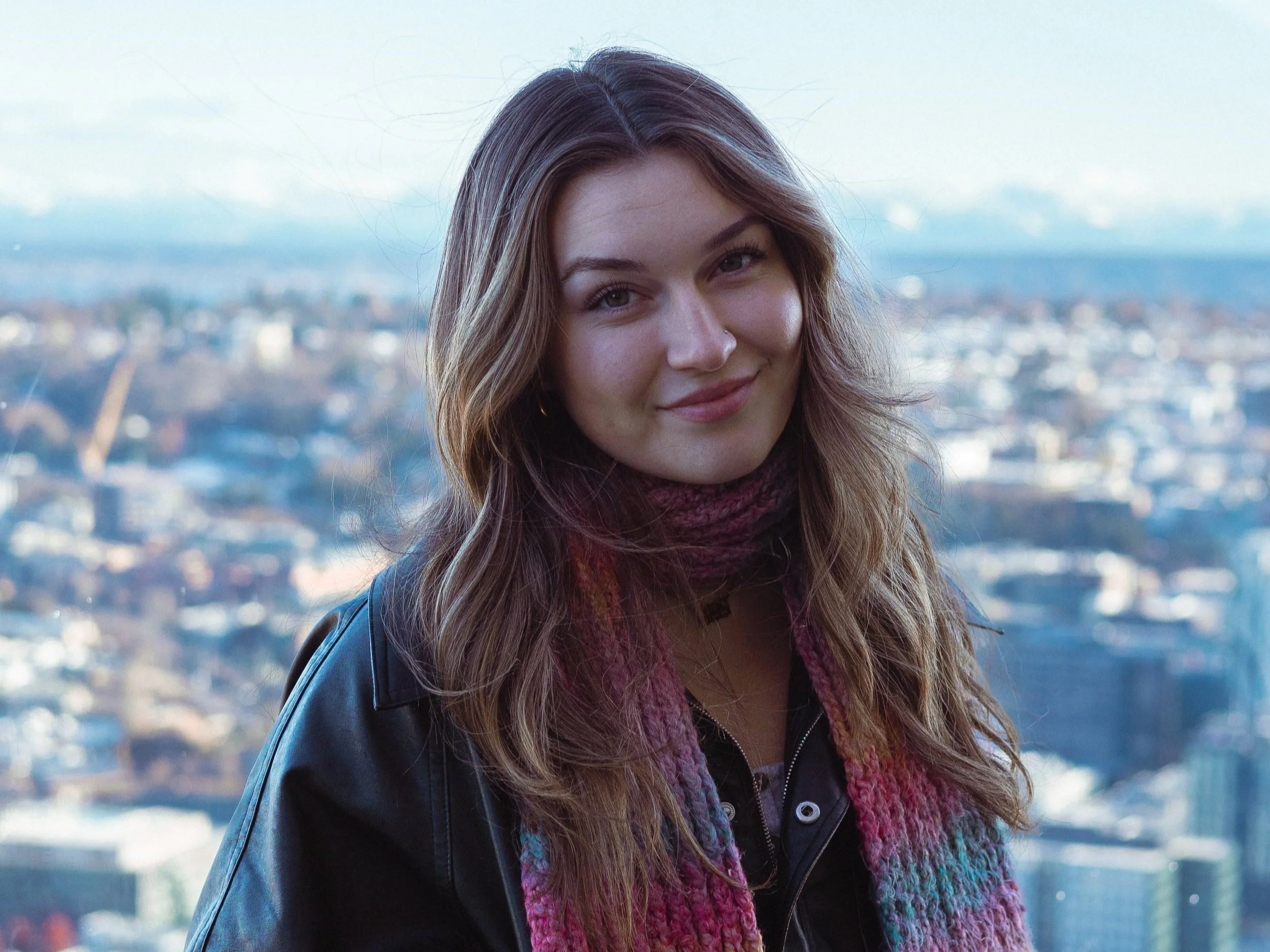 A young woman with long, wavy brown hair smiling outdoors with a cityscape and cloudy sky in the background.