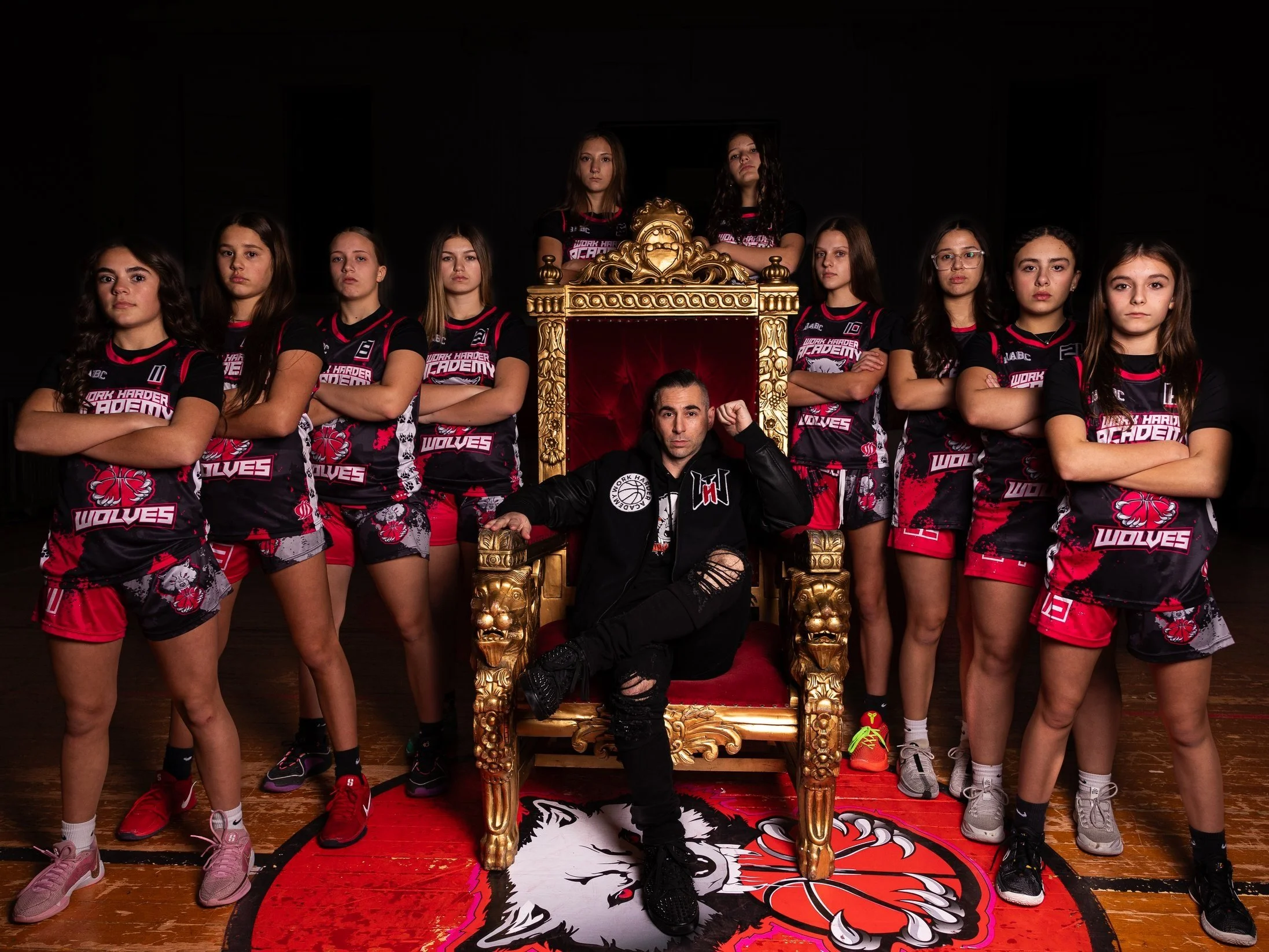 A group of female basketball players in black and red uniforms with heron team logo, stands with arms crossed around a man seated on a large ornate golden throne, in a dark room with a red lion paw rug.
