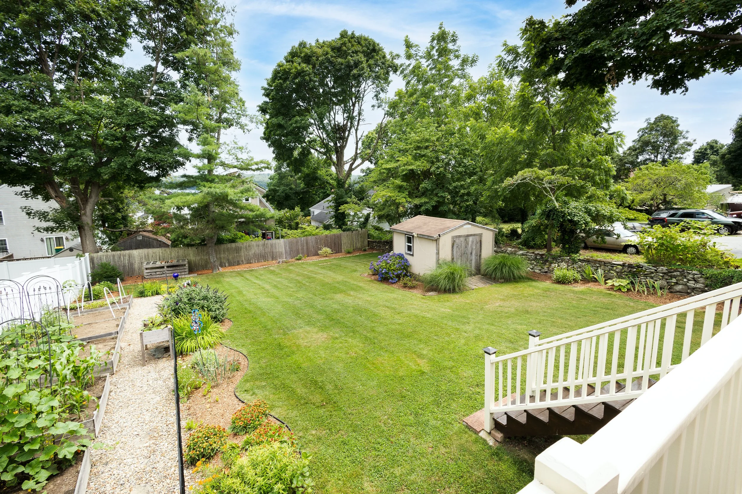 A backyard with a well-maintained green lawn, a small shed, surrounded by trees, a garden with plants and flowers, a gravel pathway, and a white railing staircase leading onto the yard.