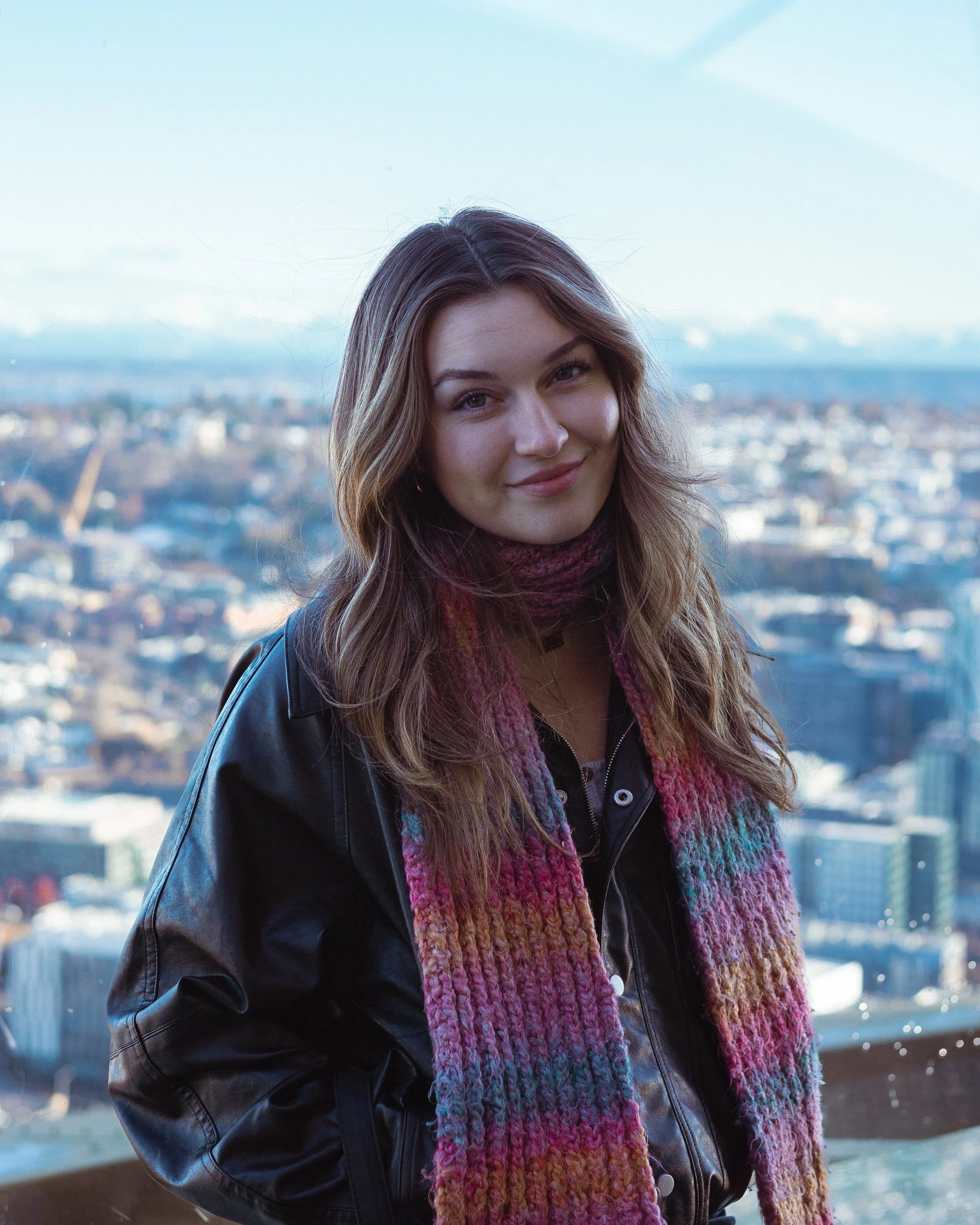 A young woman with wavy brown hair wearing a black leather jacket and colorful knitted scarf, standing outdoors with a cityscape and mountains in the background.