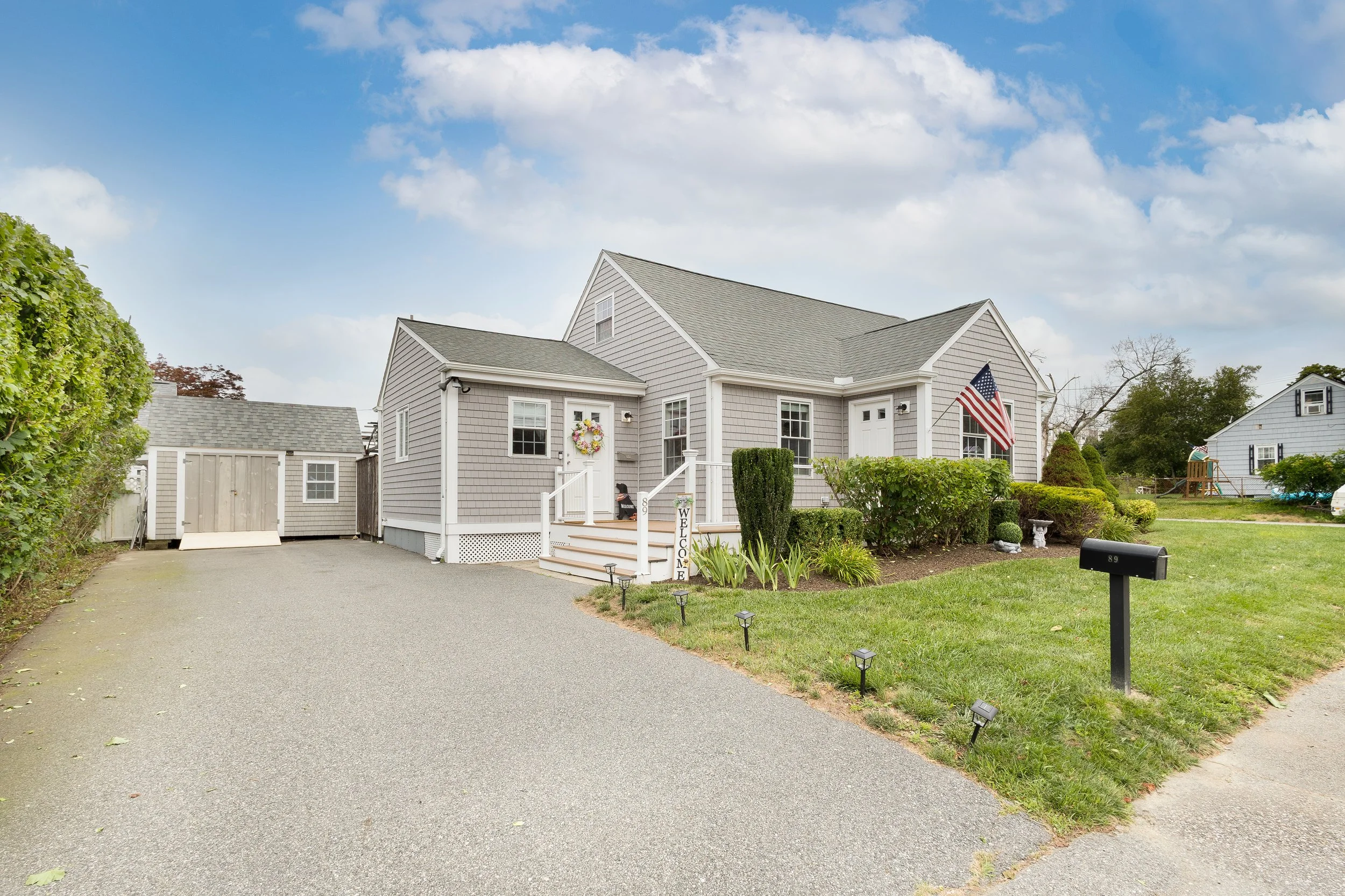 A gray single-family house with white trim, American flag, front porch, landscaped yard, black mailbox, and driveway with shed in the background on a partly cloudy day