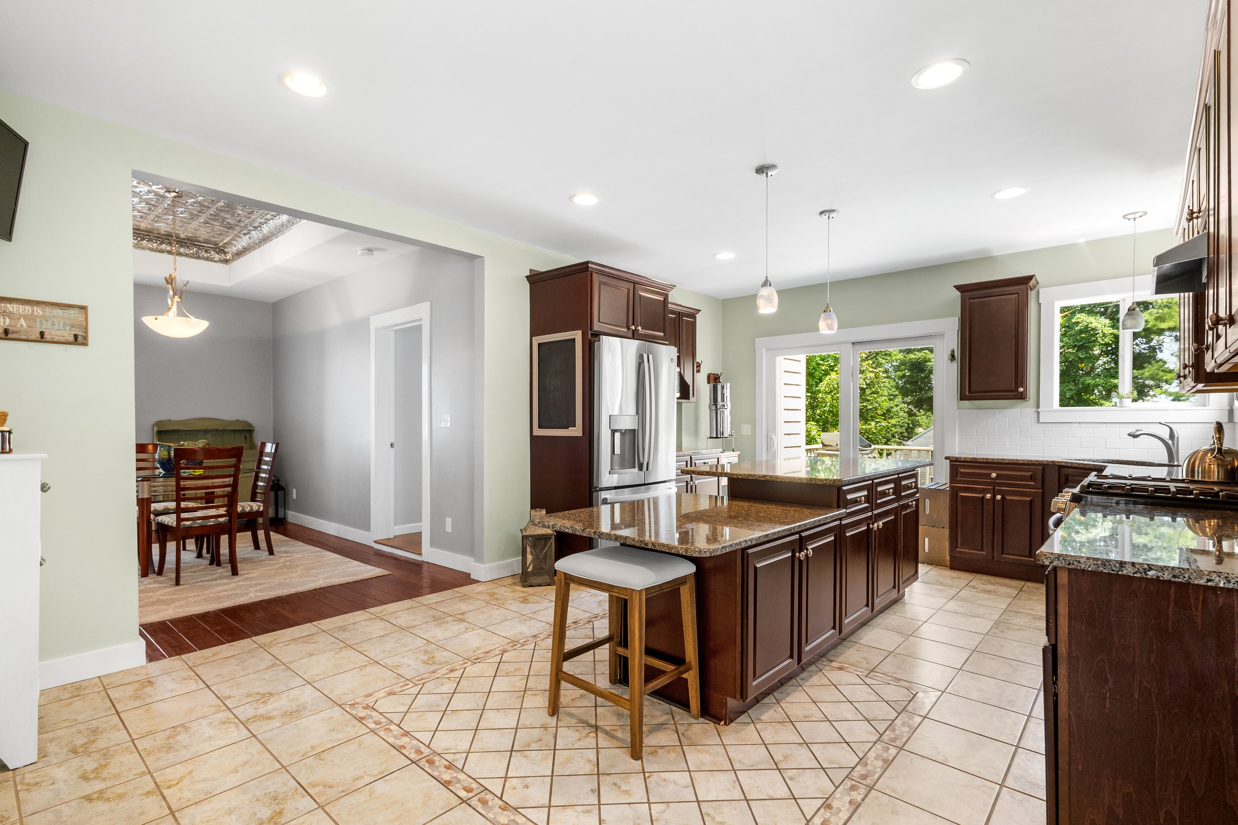 Bright kitchen with dark wood cabinets, granite countertops, and a central island with a stool. There are large windows, a sliding glass door, and ceiling lights providing natural and artificial light.