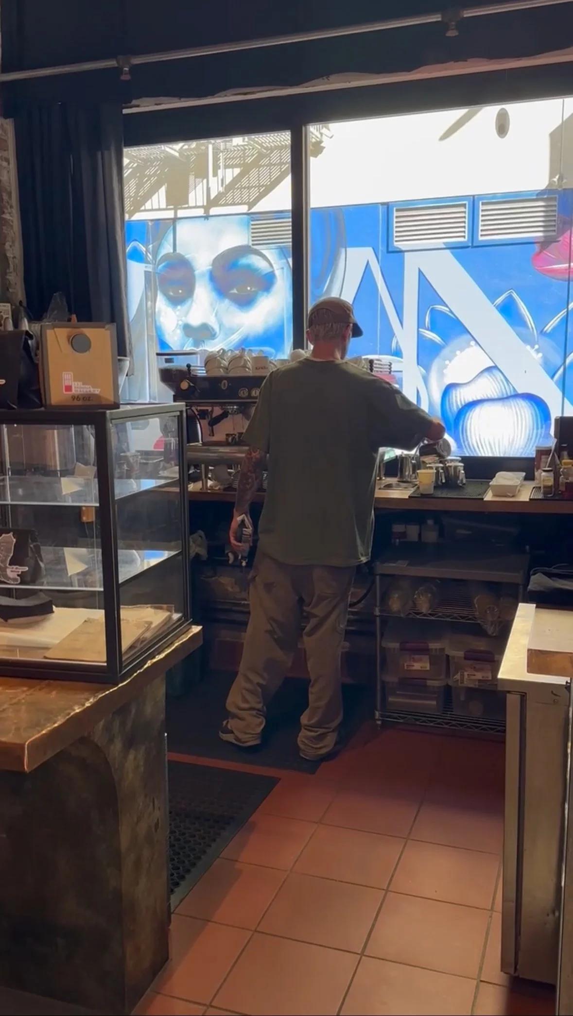 A man working behind the counter in a cafe or coffee shop with a large window showing a mural of an astronaut's face and abstract blue and white shapes outside.