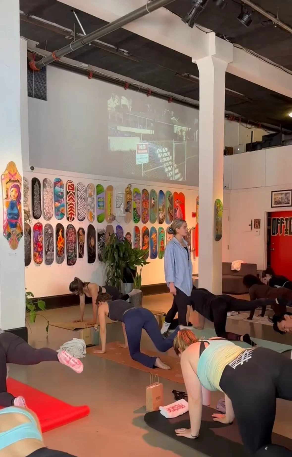People participating in a yoga class in a studio with skateboards displayed on the wall and a large projection screen on the wall.