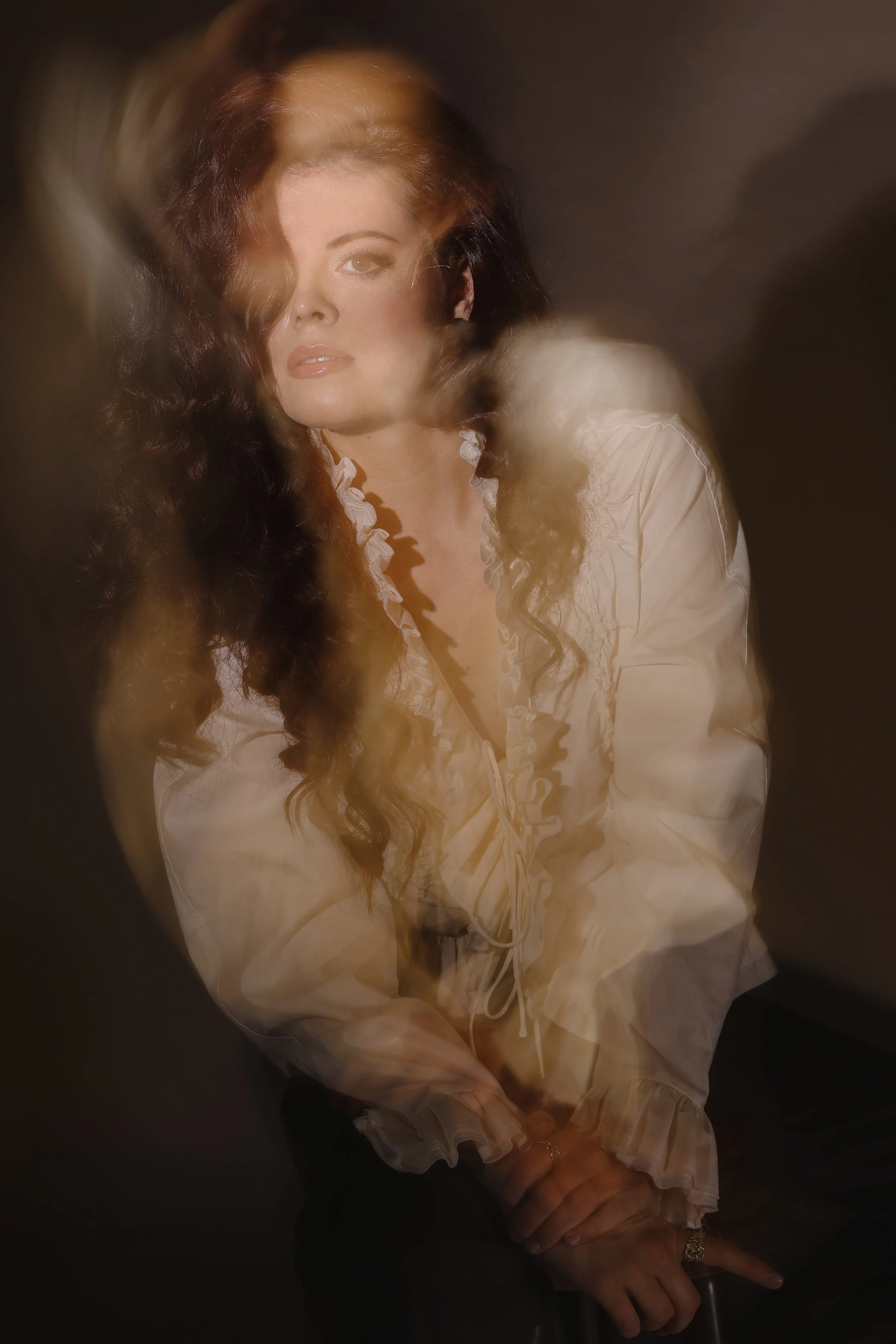 A woman with long dark hair and makeup, wearing a cream-colored blouse with ruffled details, seated with her hands crossed at the wrist, against a blurred dark background.