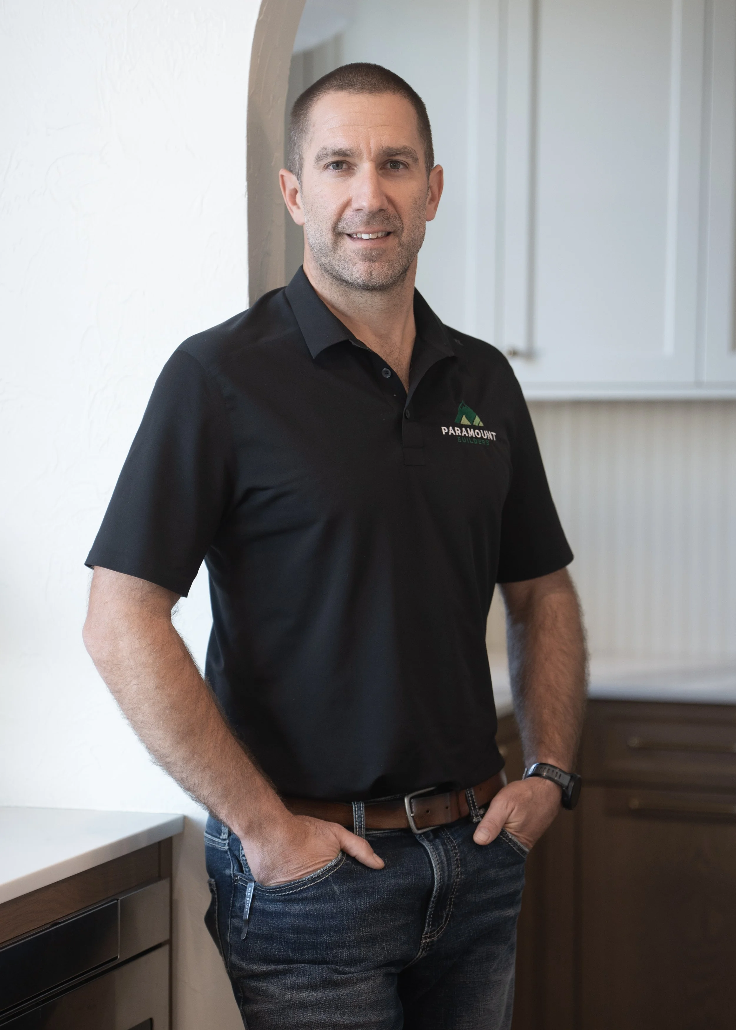 A man wearing a gray collared shirt with a logo on it, posing against a neutral background.