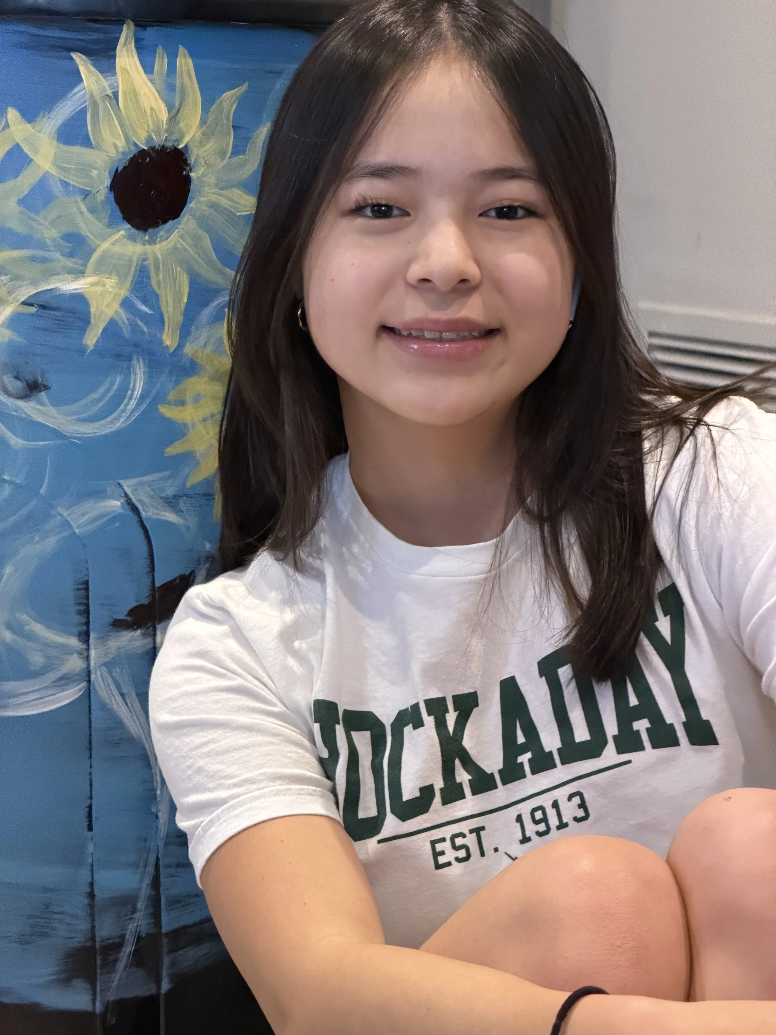 A young girl smiling while taking a selfie in front of a blue friendship-themed mural with a sunflower and water design, wearing a white T-shirt with the word 'Stockaday' and the year 1913 printed on it.