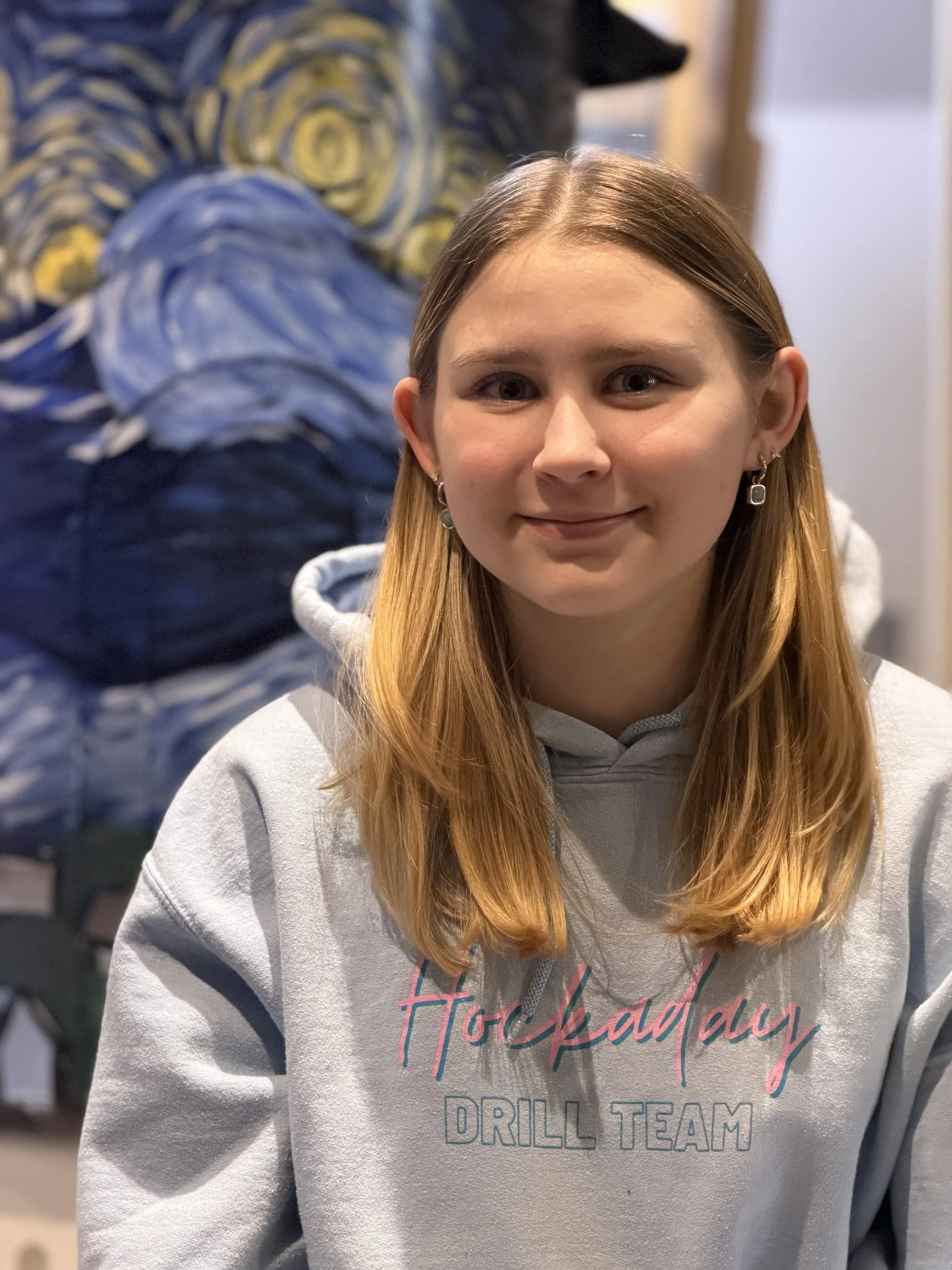 A young woman with long red hair wearing a light gray hoodie with pink and blue text, standing indoors with a decorative painting of Van Gogh's Starry Night in the background.