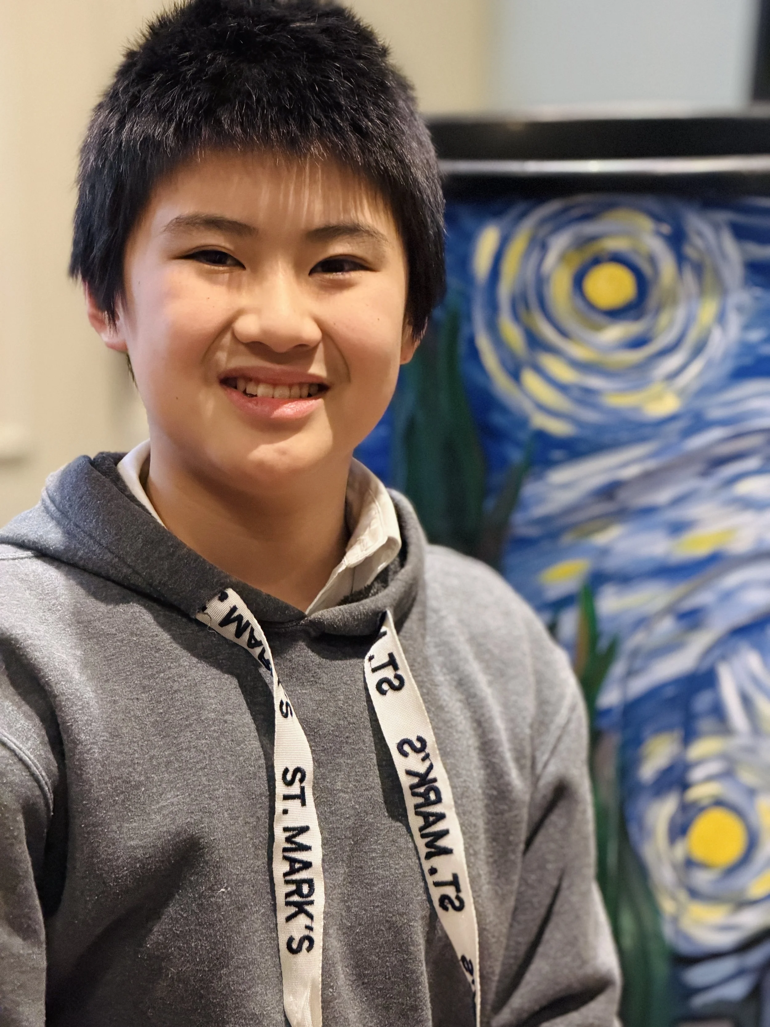Young person with short black hair smiling, wearing a gray hoodie and a white shirt, with a painting of a starry night sky in the background.