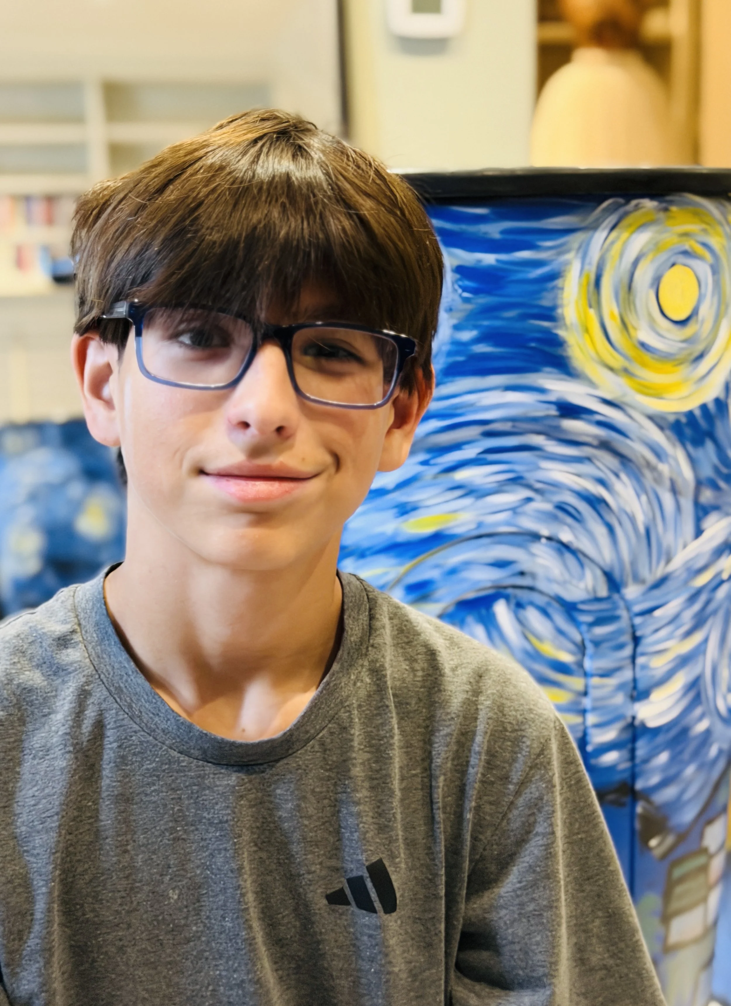 A young boy with glasses, smiling, standing in front of a vibrant painting inspired by Vincent van Gogh's Starry Night.