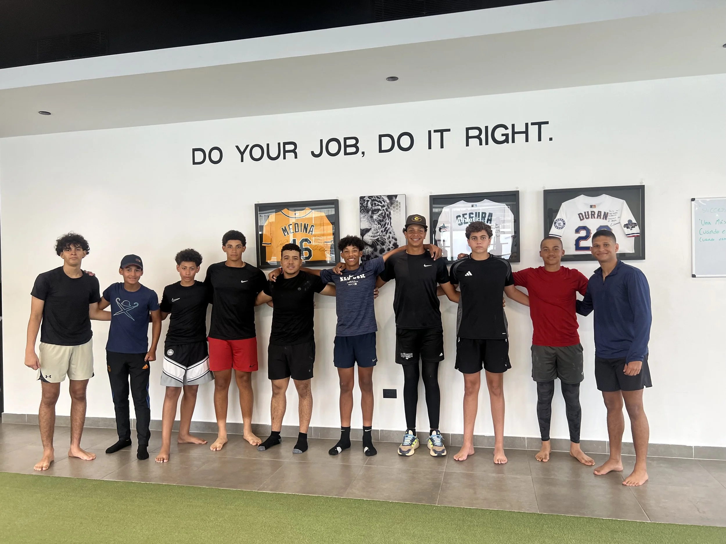 Group of nine young men standing side by side with arms around each other's shoulders in front of a wall with framed sports jerseys and a tiger picture. The wall has the phrase 'Do your job, do it right' written on it.