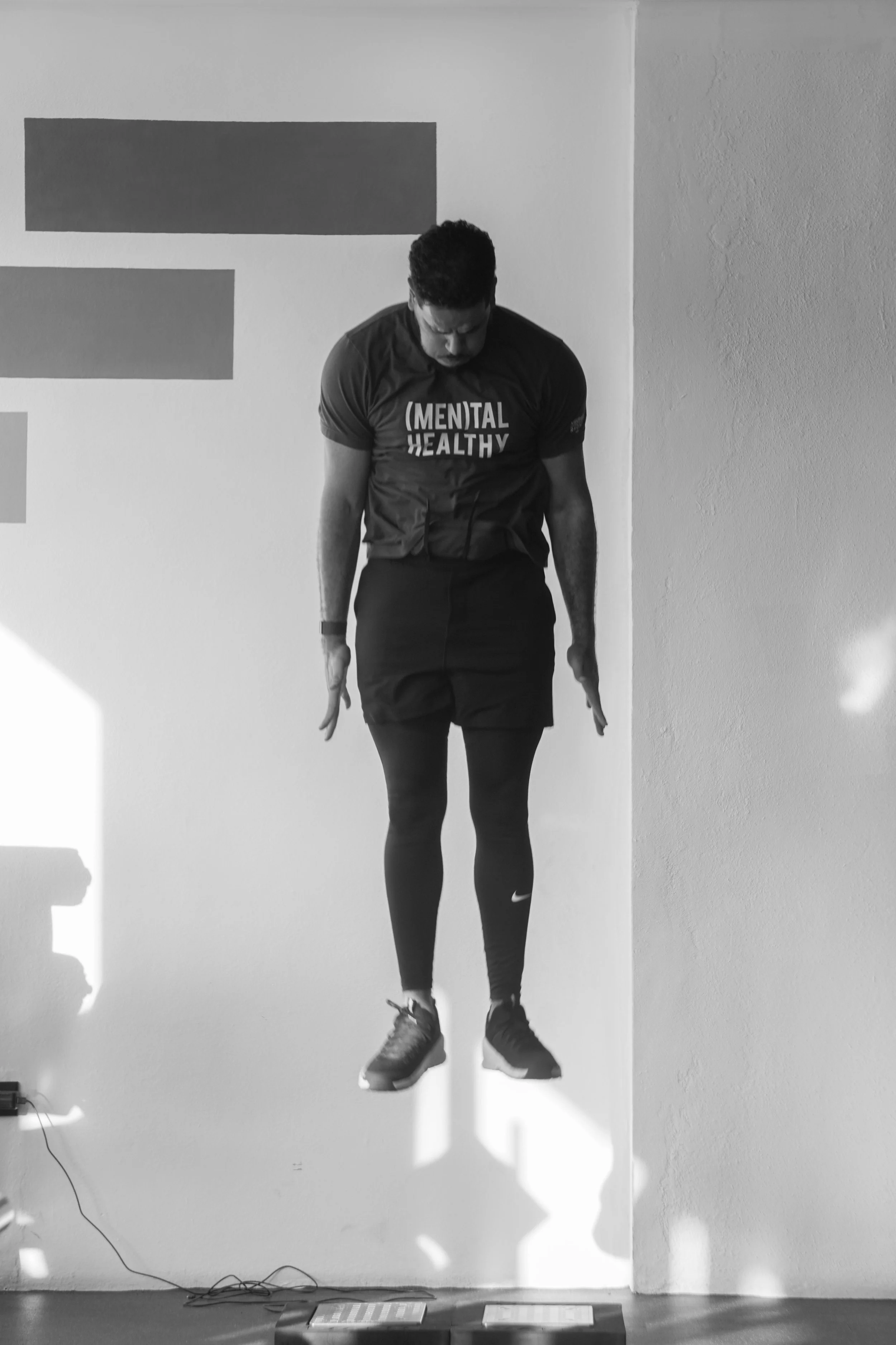 A man in athletic clothing is doing a jump exercise against a wall. He is wearing a t-shirt that reads '(MENTAL) HEALTHY', shorts, and athletic shoes.