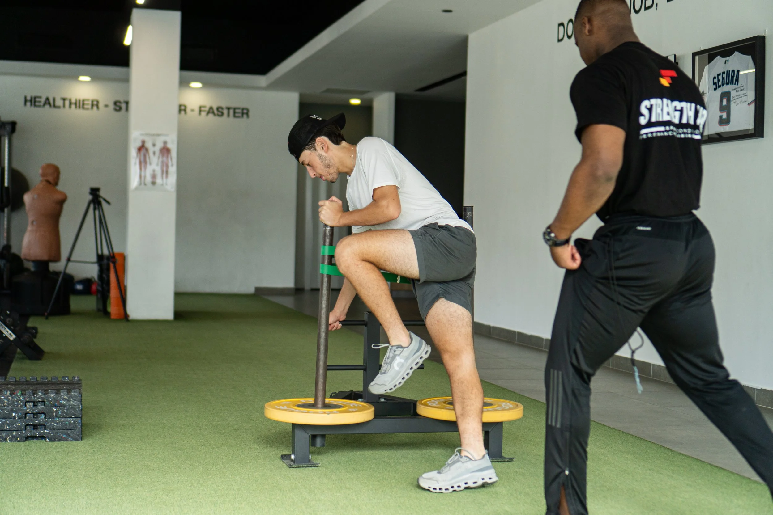 Young man performing a step-up exercise on a platform with a coach nearby, inside a gym with fitness equipment and motivational signs.