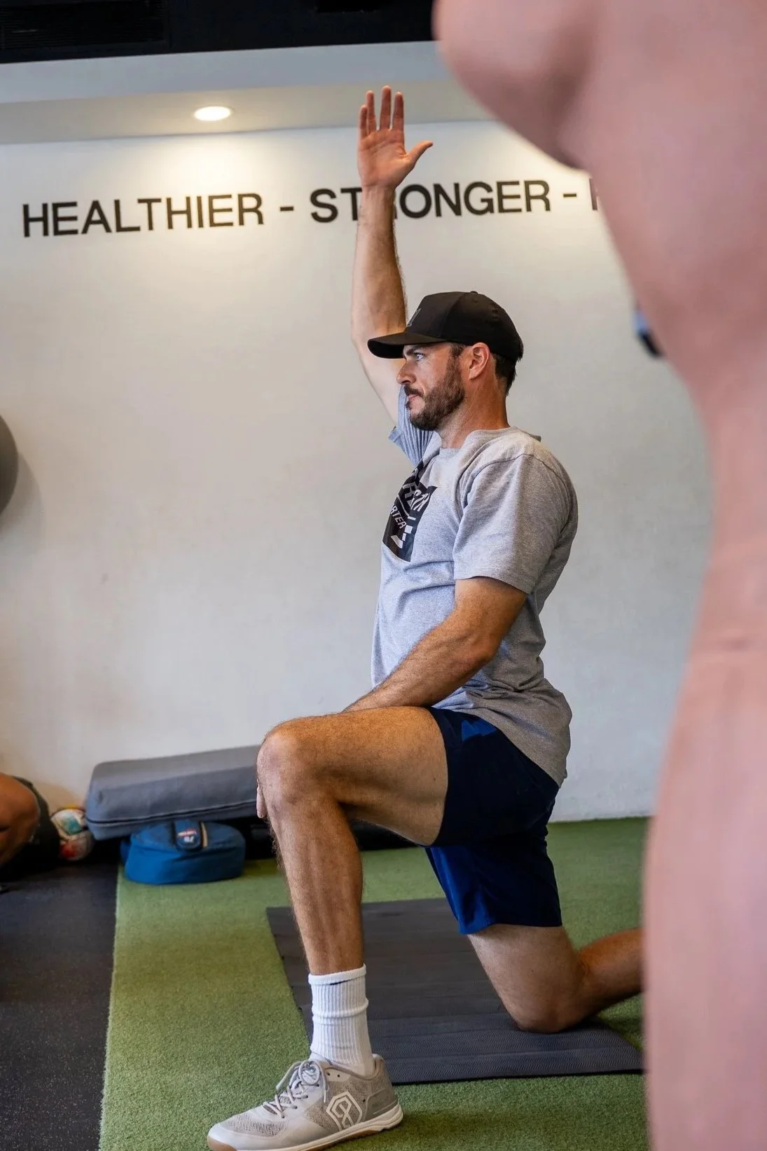 A man in a gray t-shirt, black shorts, white sneakers, and a black cap is performing a kneeling exercise with one knee on a gym mat, raising one arm while focusing straight ahead. Other people and gym equipment are visible in the background, and a sign on the wall reads 'HEALTHIER - STRONGER -'