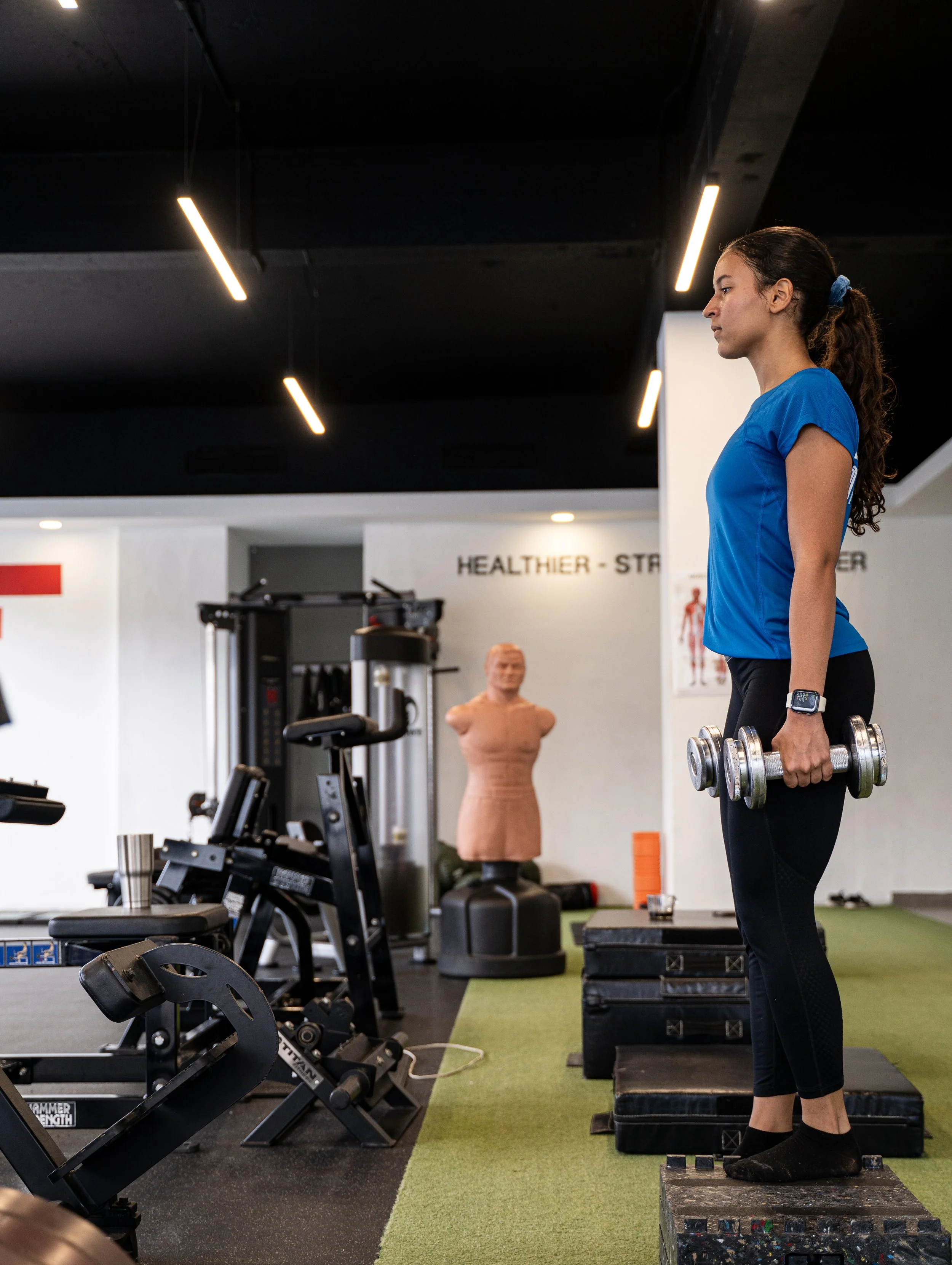 Woman in blue shirt lifting dumbbells in a gym with fitness equipment and a training mannequin in the background.