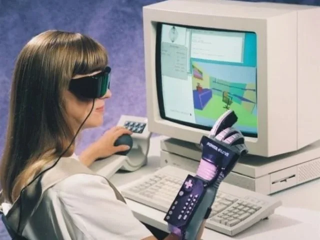 Woman wearing a virtual reality headset and gloves using a computer with a 3D modeling program on the screen.