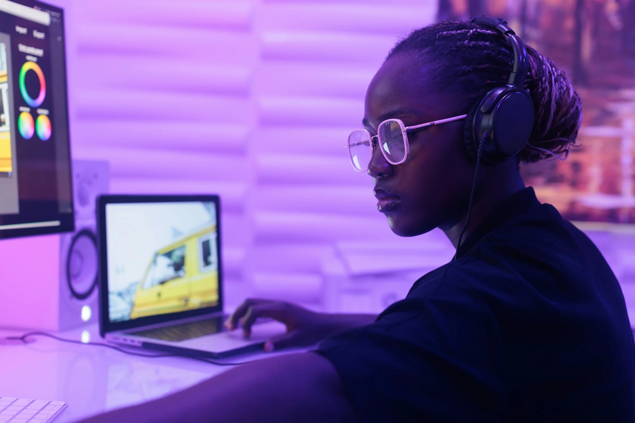 A woman with glasses and braided hair working on a laptop at a desk with multiple computer screens in a room with purple ambient lighting.
