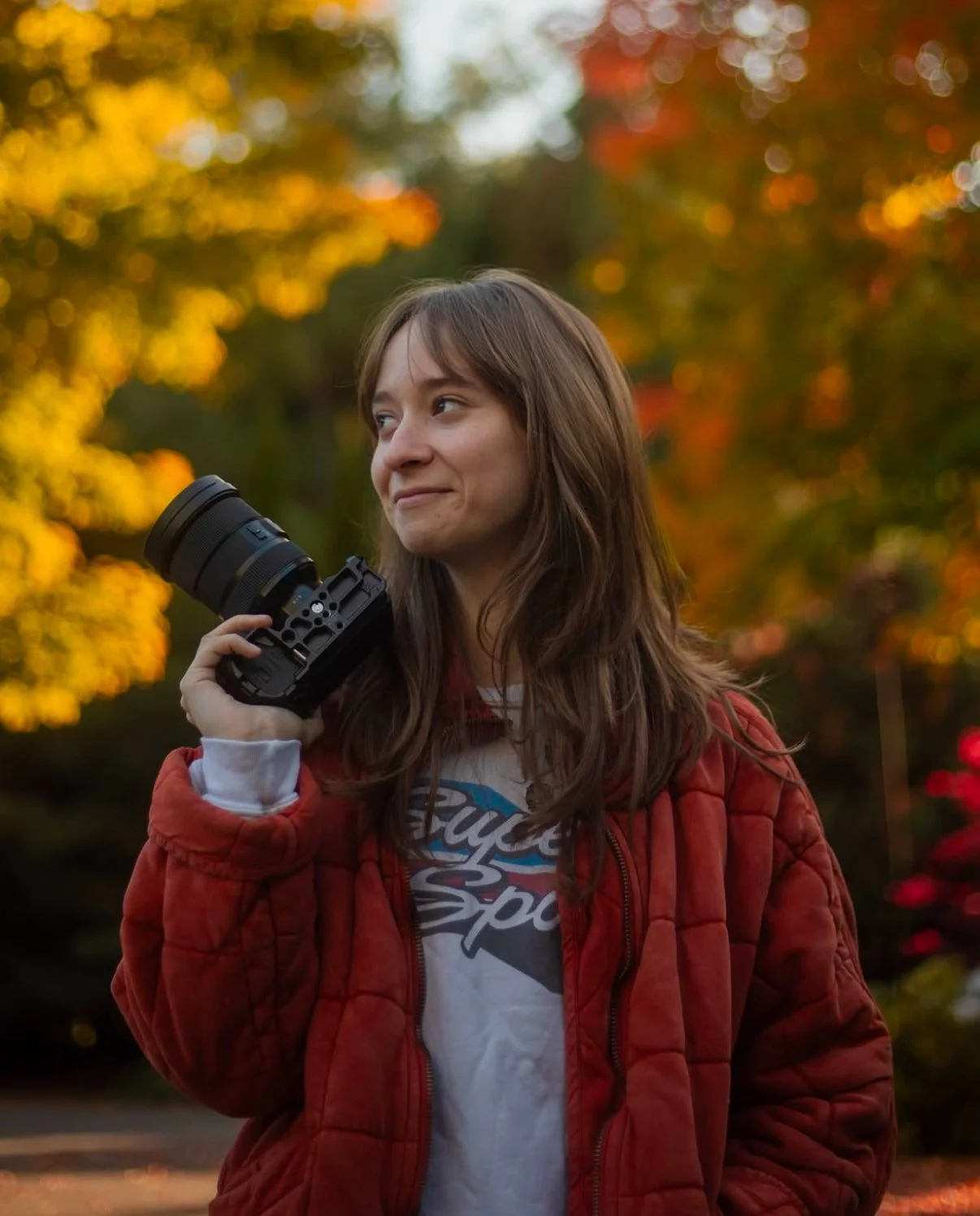 A young woman with long brown hair, wearing a red quilted jacket and a vintage t-shirt, is standing outdoors during fall and holding a professional camera with a large lens on her shoulder. She is smiling slightly and looking to the side, with colorful autumn leaves in the blurred background.