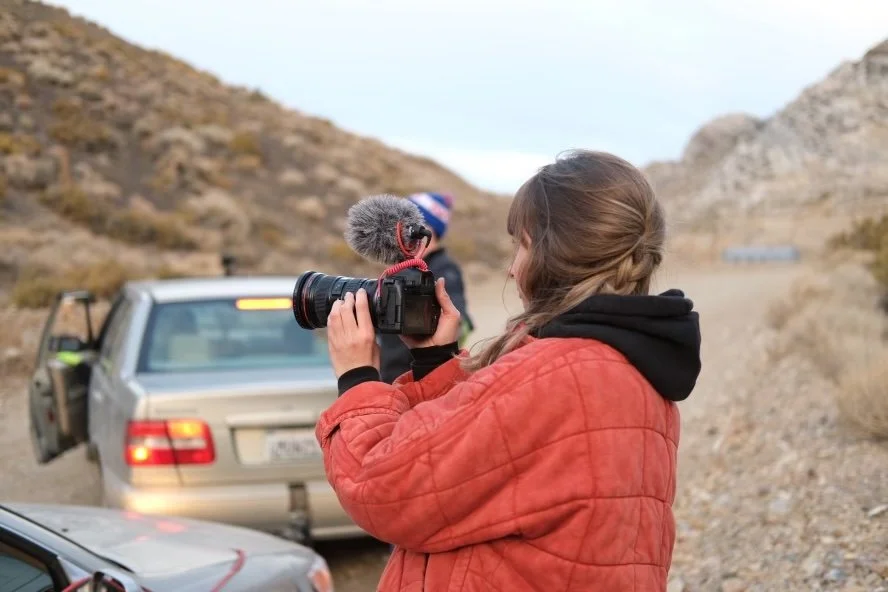A woman with brown hair, wearing an orange jacket, is holding a camera with a microphone, filming in a rocky outdoor landscape. There are two cars behind her, one with the door open, and a person in a hat in the background.