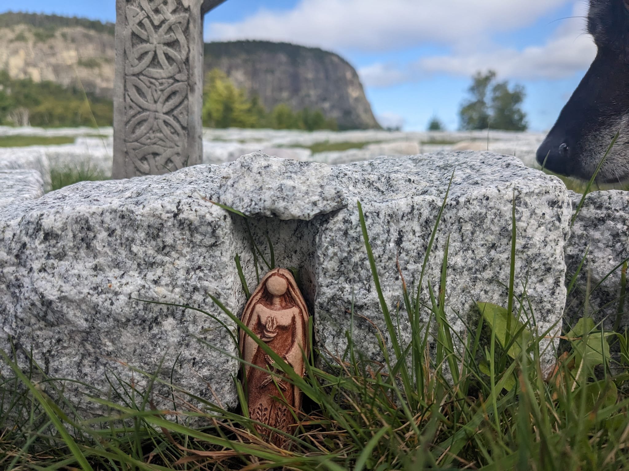 A carved icon of brigid standing upright on a rocky ground outdoors, with a large black dog laying nearby. The background shows a grassy area, trees, a large cliff or mountain, and a partly cloudy sky.