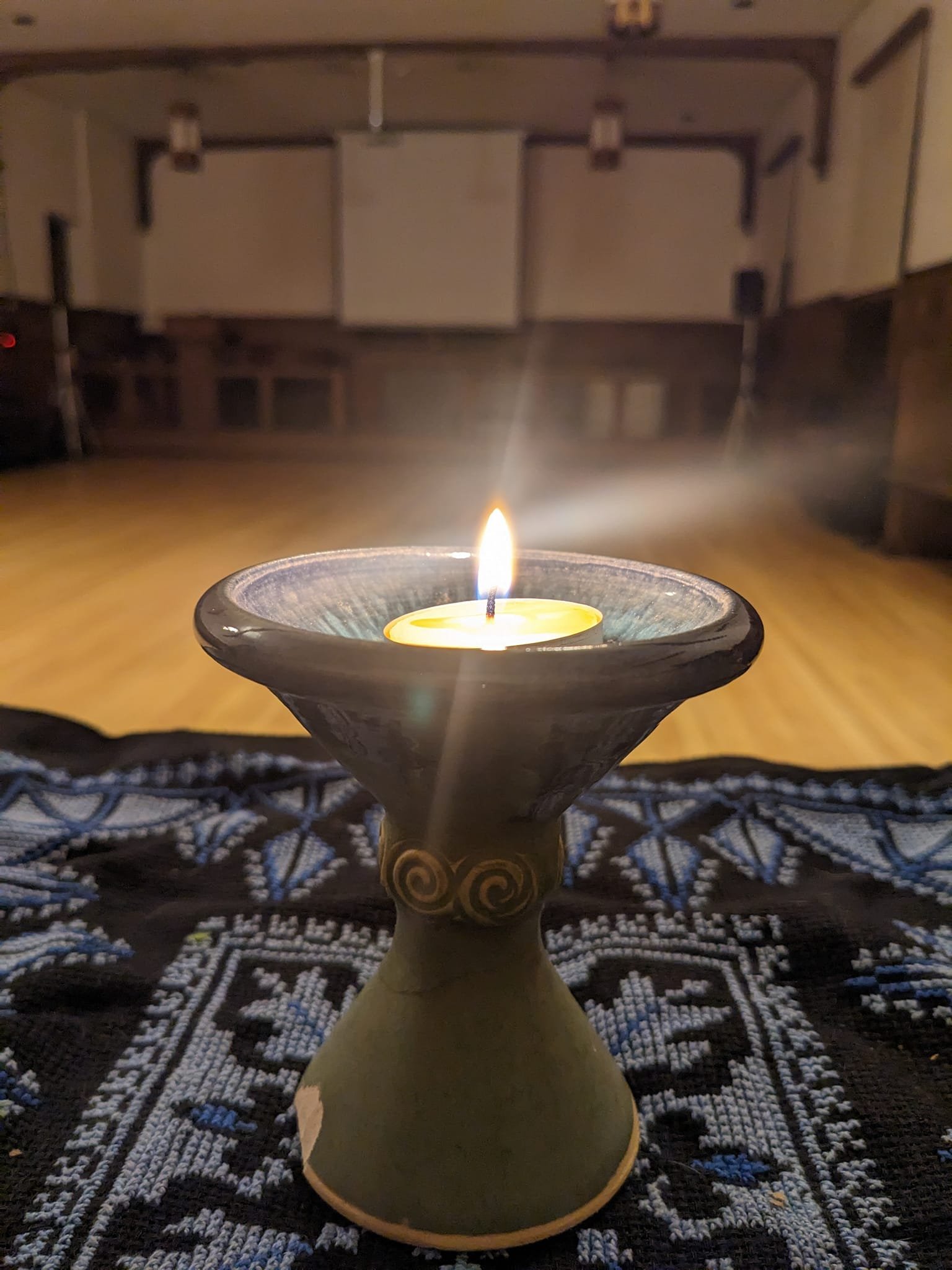 A lit candle on a decorative candle holder placed on an altar cloth, with a blurred background of a room with some furniture and a white ceiling.