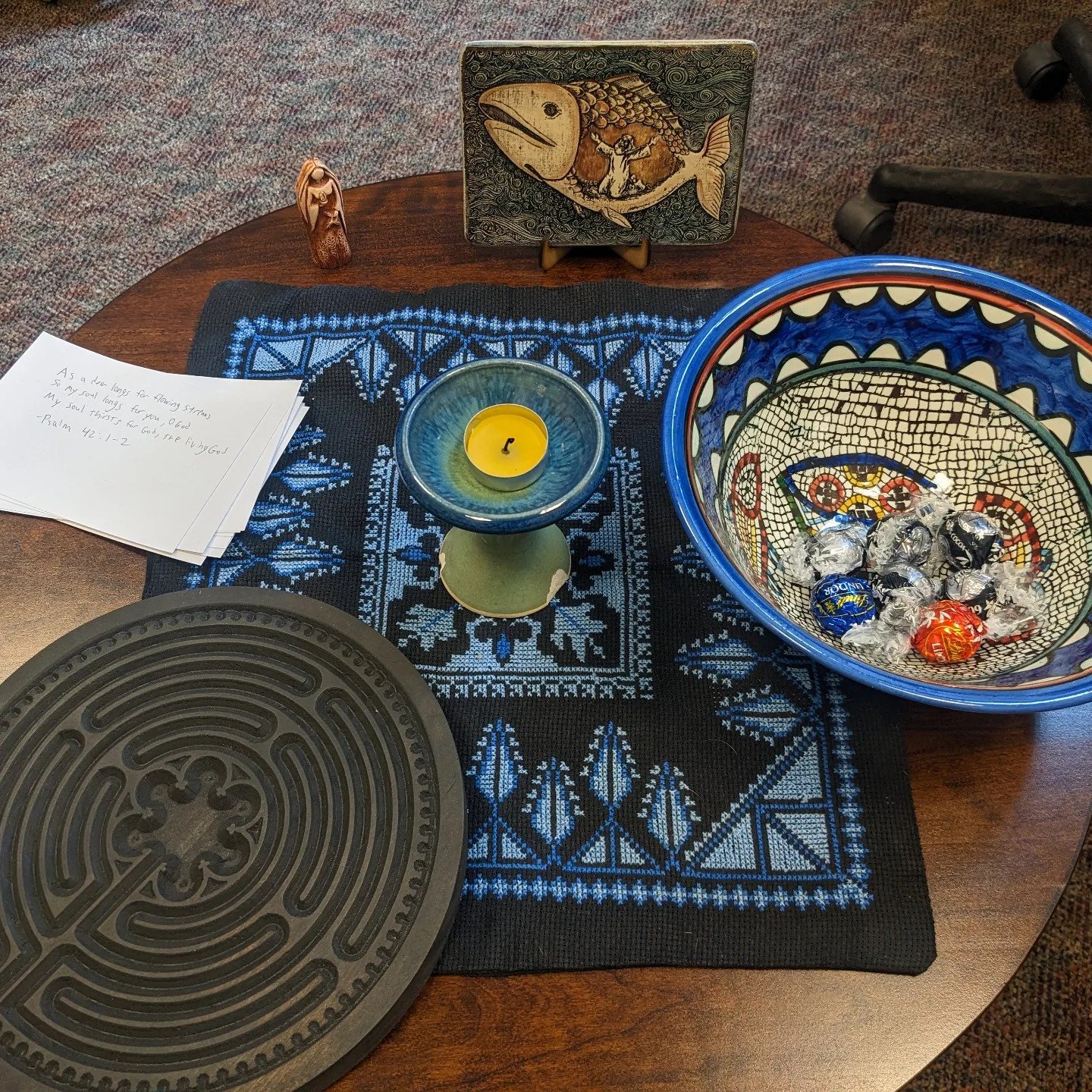 A table decorated with a black and blue tablecloth, holding a yellow candle, a blue decorative plate, a black round tray, a note with handwritten Bible verse, a small wooden sculpture, a framed picture of a fish with a man, and a colorful bowl filled with wrapped candies.