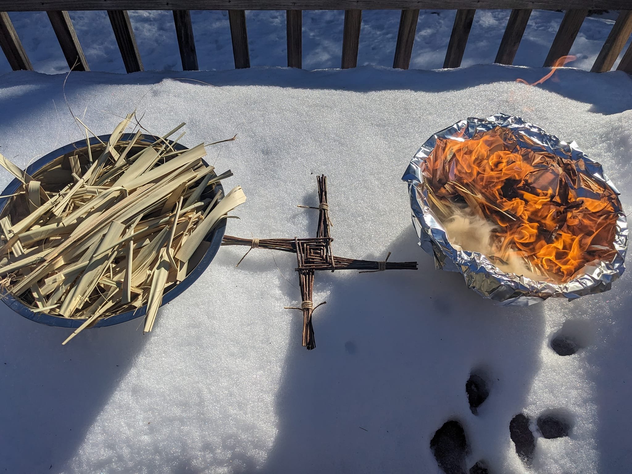 Snow-covered surface with a small campfire in aluminum foil and a bundle of dried grass on the left, with a wooden cross in the center, and footprints in the snow.