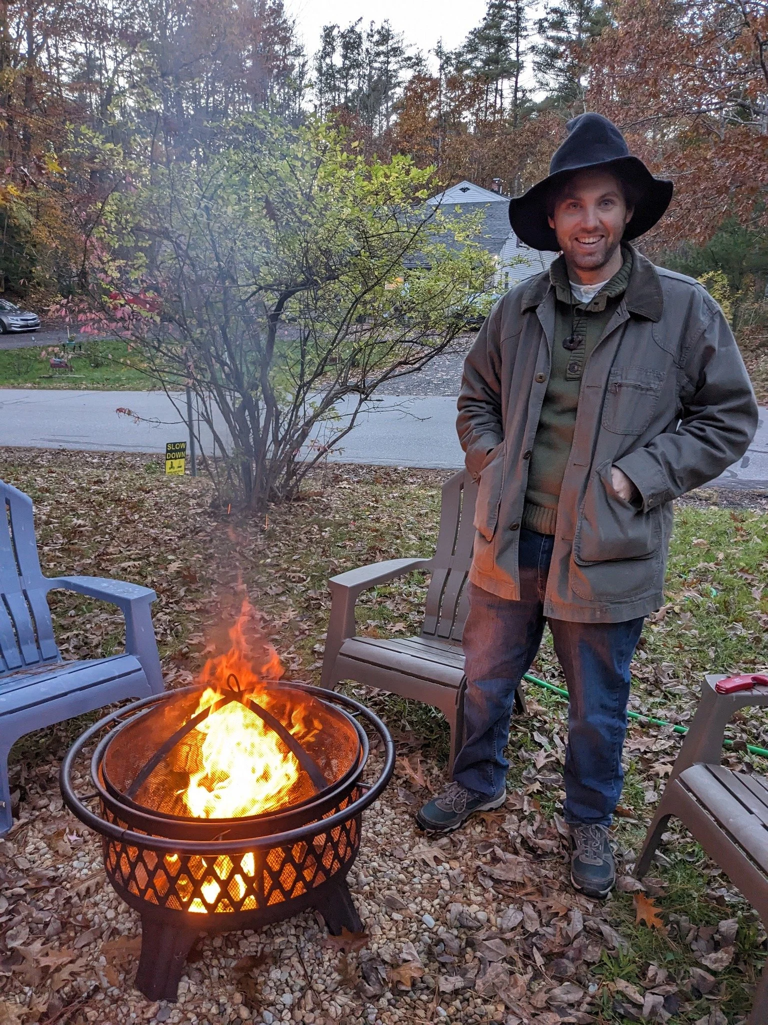 A smiling person wearing a black hat and outdoor clothing standing outdoors near a fire pit with a fire, with chairs around and trees in the background, during autumn.
