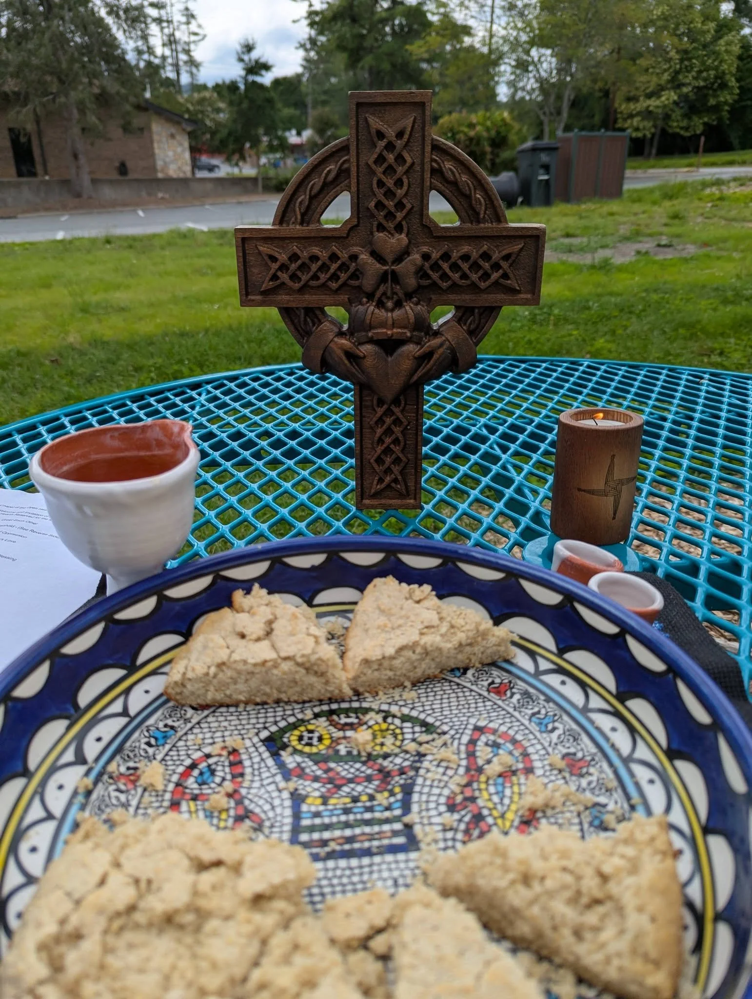A decorative wooden Celtic cross on a metal table outdoors. A ceramic cup and a small lit candle are nearby, with green grass, trees, and residential houses in the background.