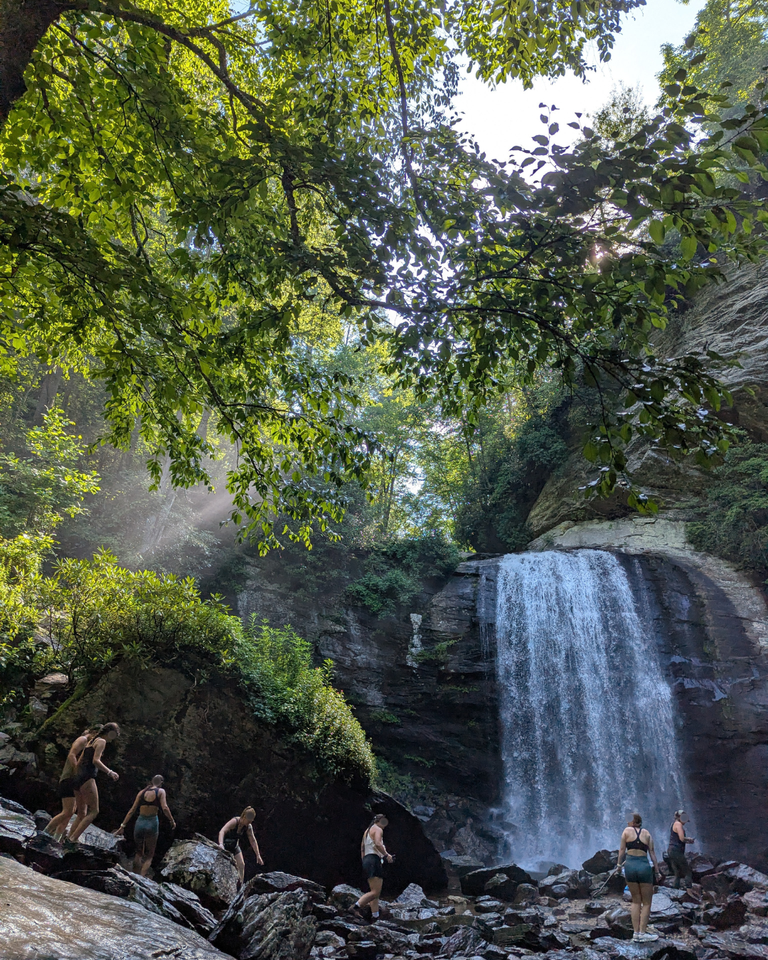 Group of people hiking along rocky terrain near a waterfall in a lush forest with sunlight filtering through green leaves.