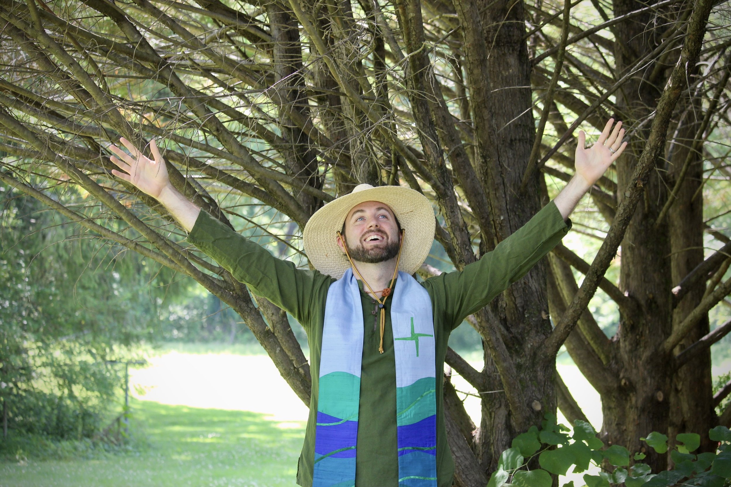 A man wearing a wide-brimmed straw hat, green shirt, and a light blue stole with green crosses, standing outdoors with arms raised in front of a tree with many branches and leaves, in a lush green park.