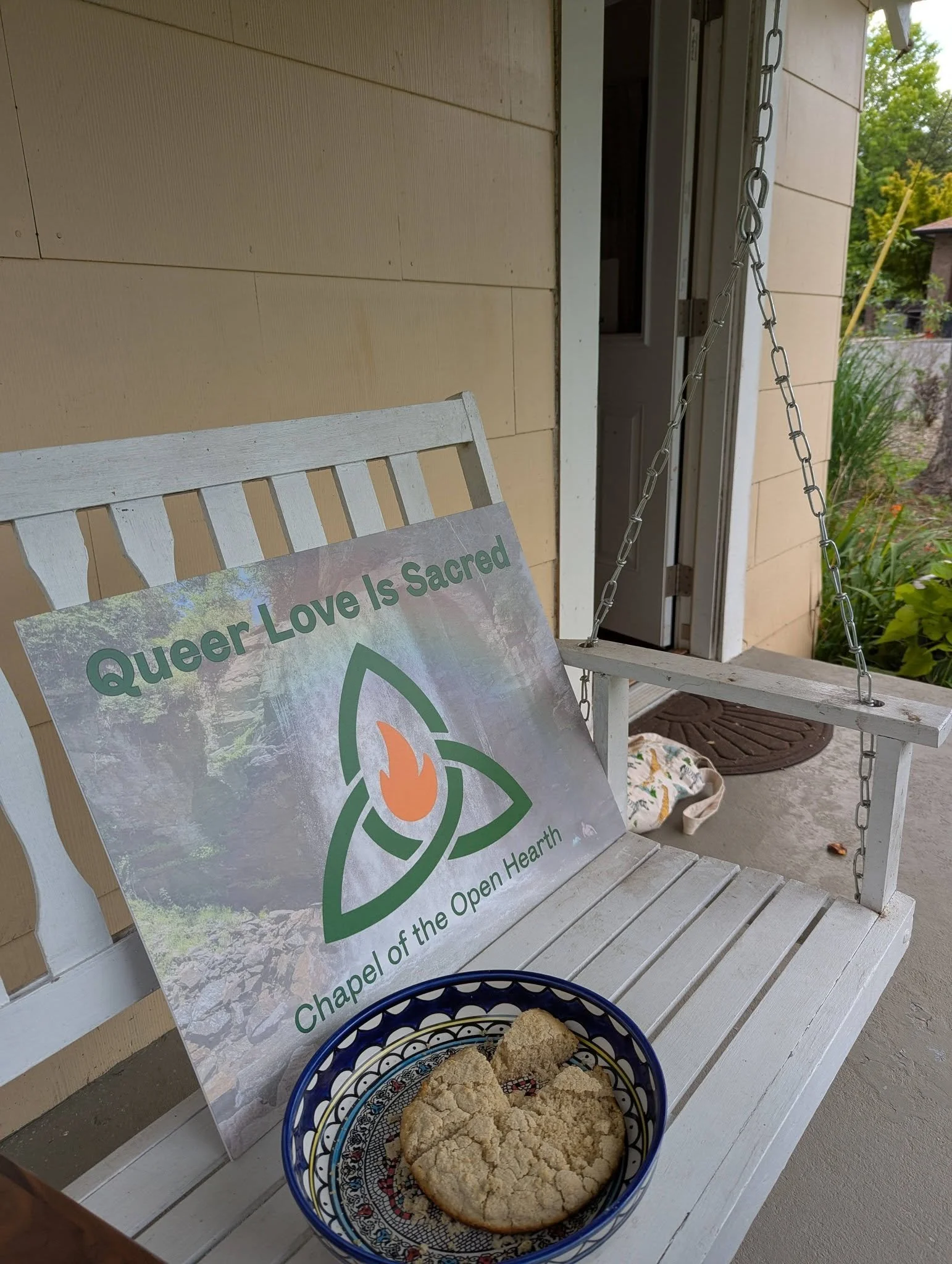A white wooden bench with a sign that reads 'Queer Love Is Sacred' leans against the backrest. Next to the bench, there is a metal chain suspended from the wall, connected to a saddle-like seat. In front of the bench, a blue bowl with a colorful pattern contains large, irregularly shaped pieces of bread or baked goods. The setting appears to be on a porch with a brick wall and greenery in the background.